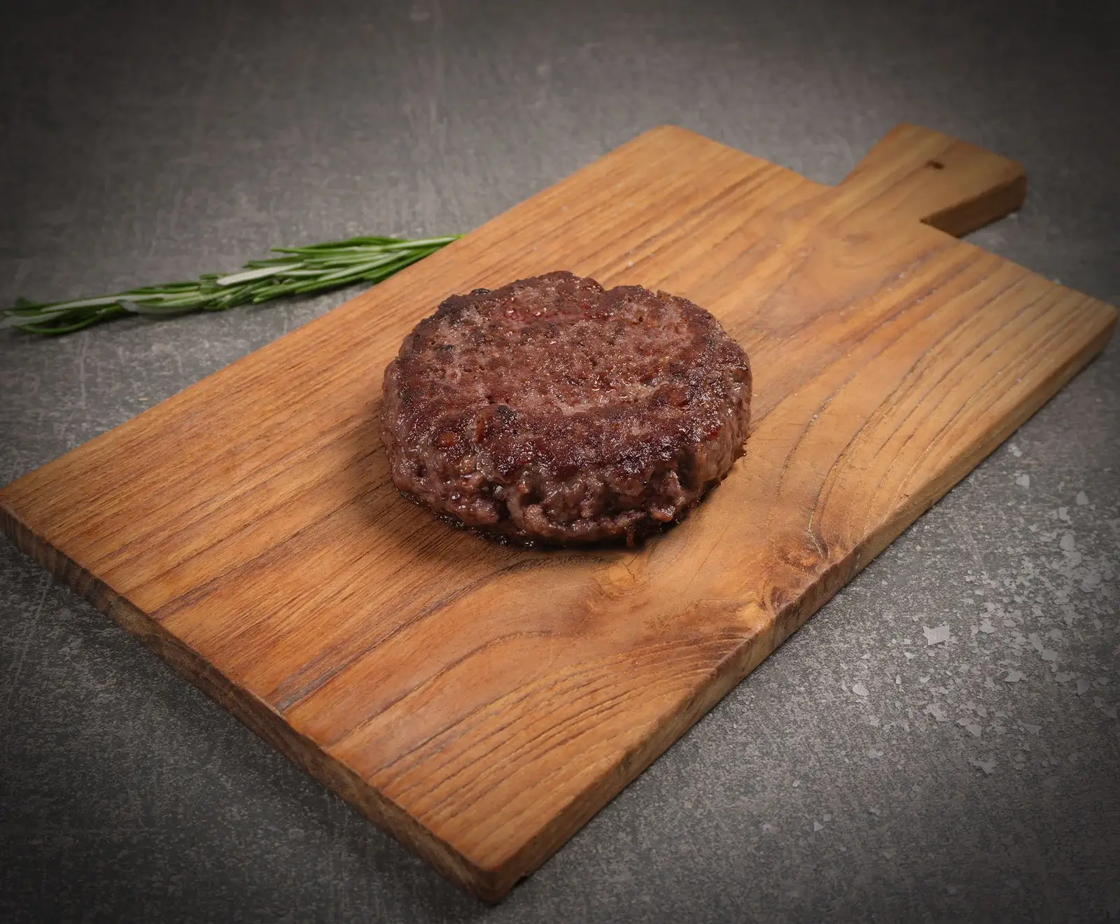 Ein saftiges, gebratenes Burger-Patty liegt auf einem Holzschneidebrett. Im Hintergrund ist ein Rosmarinzweig auf dunklem Untergrund zu sehen.