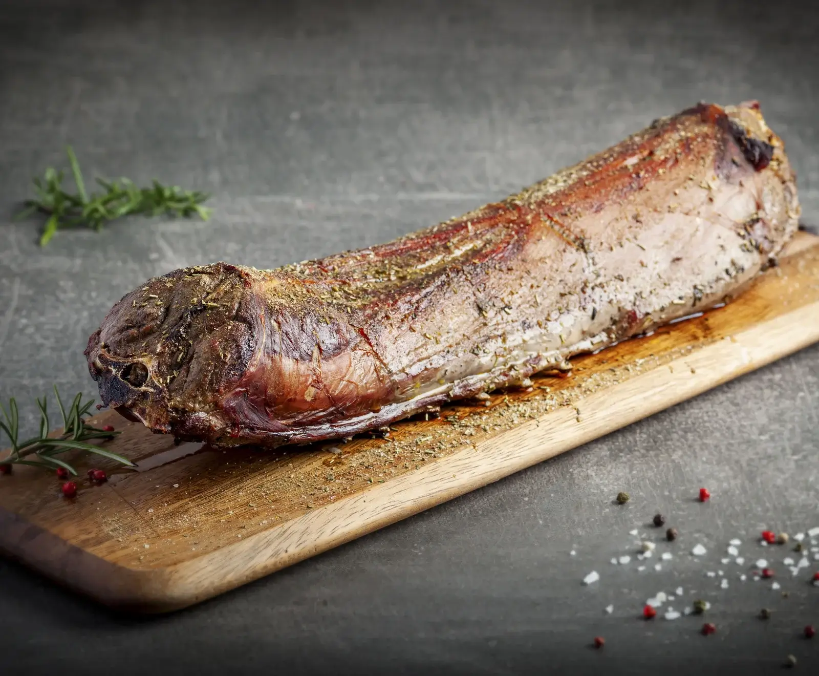 Ein goldbraun gebratener, mit Kräutern und Gewürzen bestreuter Fleischbraten liegt auf einem Holzschneidebrett. Rosmarinzweige, Pfefferkörner und grobes Salz sind auf dem dunklen Hintergrund verteilt.