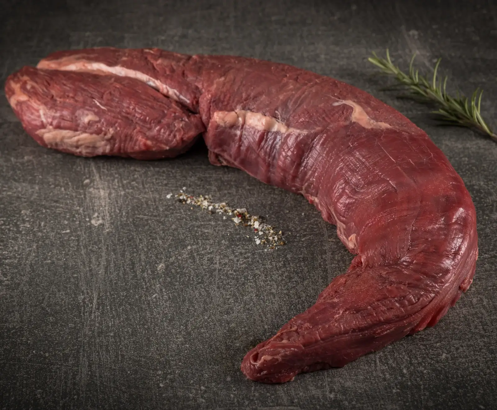 Ein ganzes, rohes Rinderfilet liegt auf einer dunklen, strukturierten Oberfläche, begleitet von grobem Salz, Pfeffer und einem Rosmarinzweig.