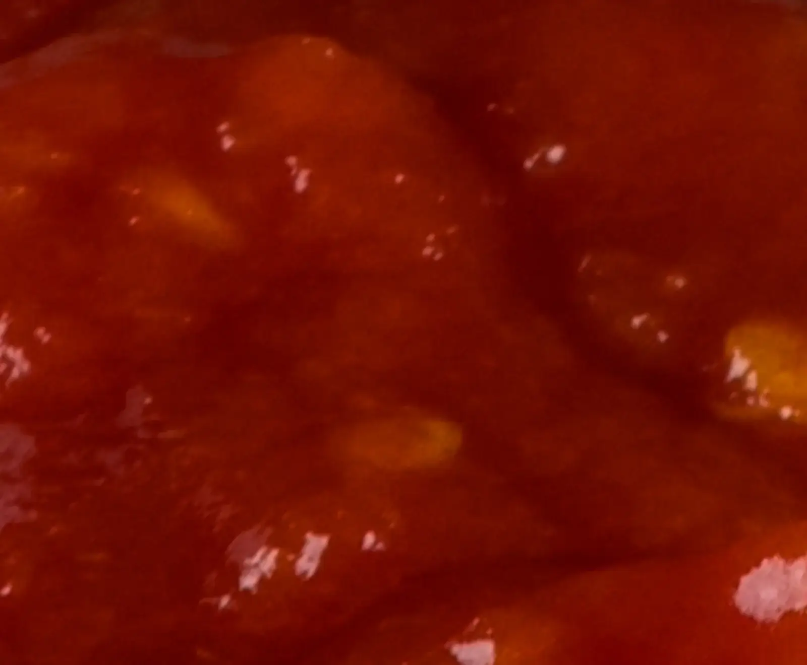 Nahaufnahme einer leuchtend roten, dickflüssigen Sauce mit kleinen, helleren, gelblichen Stücken, die an Frucht oder Gemüse erinnern.