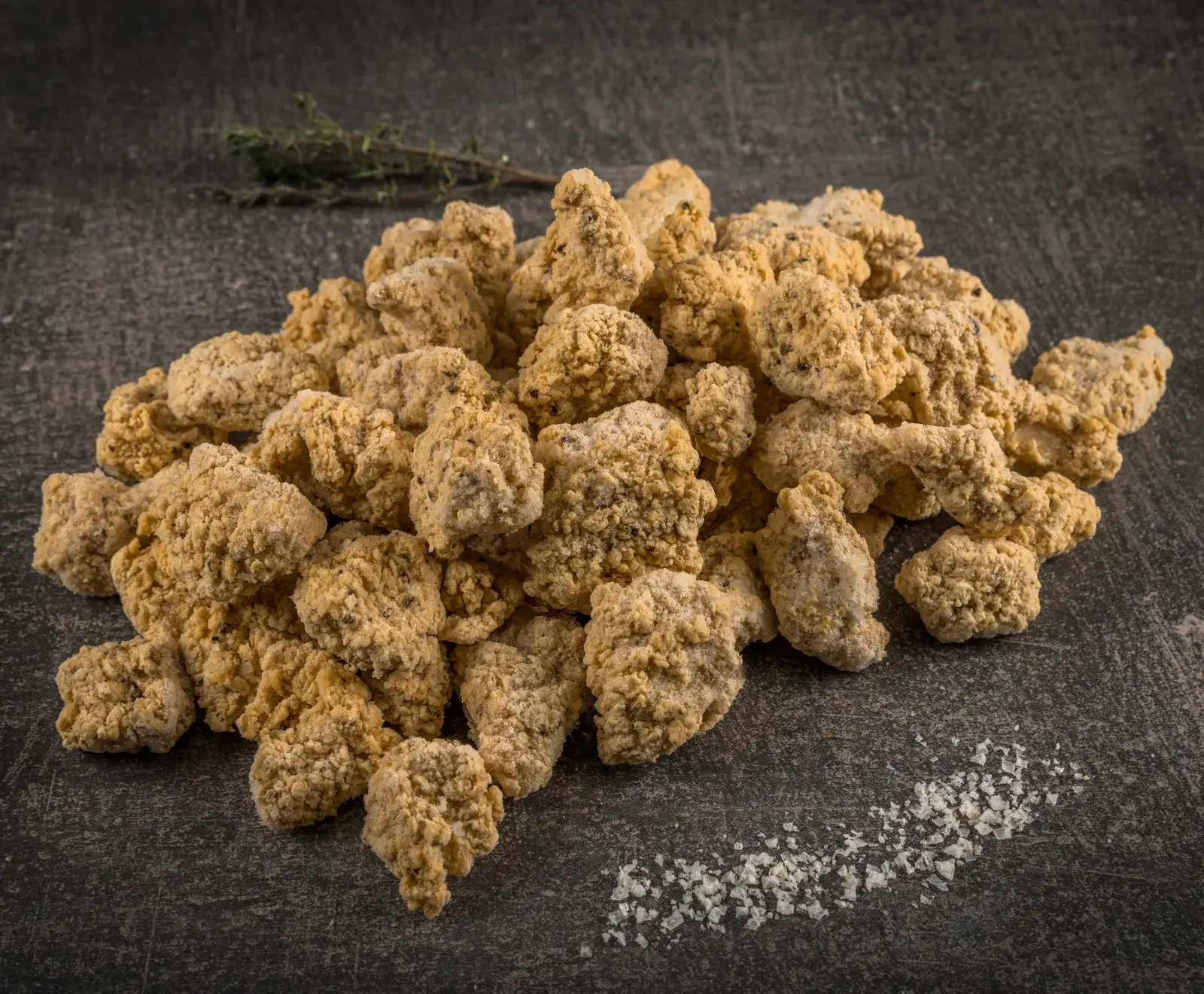 Ein Haufen gefrorener, panierter Hähnchenstücke auf dunklem Untergrund, garniert mit einem Thymianzweig und groben Salzkristallen.