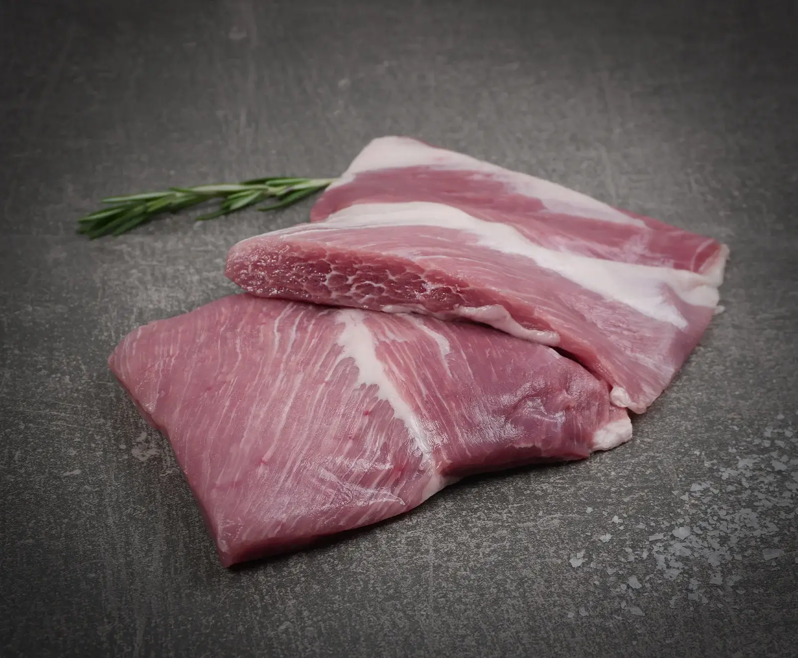 Zwei rohe Stücke Schweinebauch mit mageren und fetten Schichten liegen auf einer dunklen, strukturierten Oberfläche. Ein Rosmarinzweig und grobes Salz sind im Hintergrund zu sehen.
