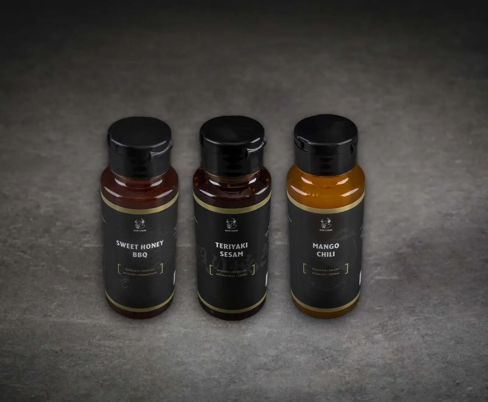 Drei Flaschen Don Carne Saucen/Marinaden stehen auf einer dunklen Oberfläche. Von links nach rechts: Sweet Honey BBQ (dunkelrotbraun), Teriyaki Sesam (dunkelbraun) und Mango Chili (orange-gelb), alle mit schwarzen Etiketten und goldenen Akzenten.