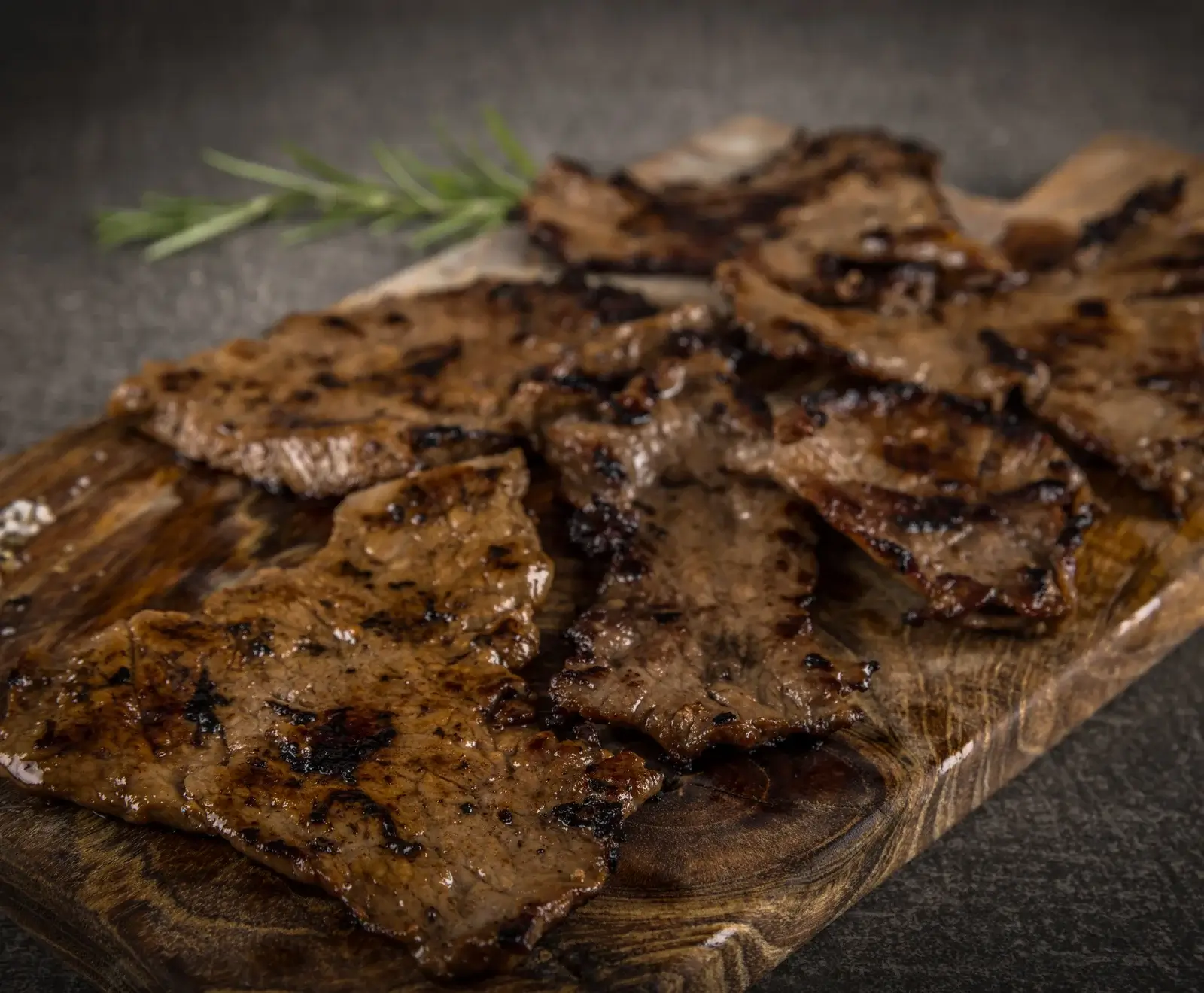 Dünne, gegrillte Rinderstreifen mit Grillspuren auf einem dunklen Holzschneidebrett, mit Rosmarin im Hintergrund.