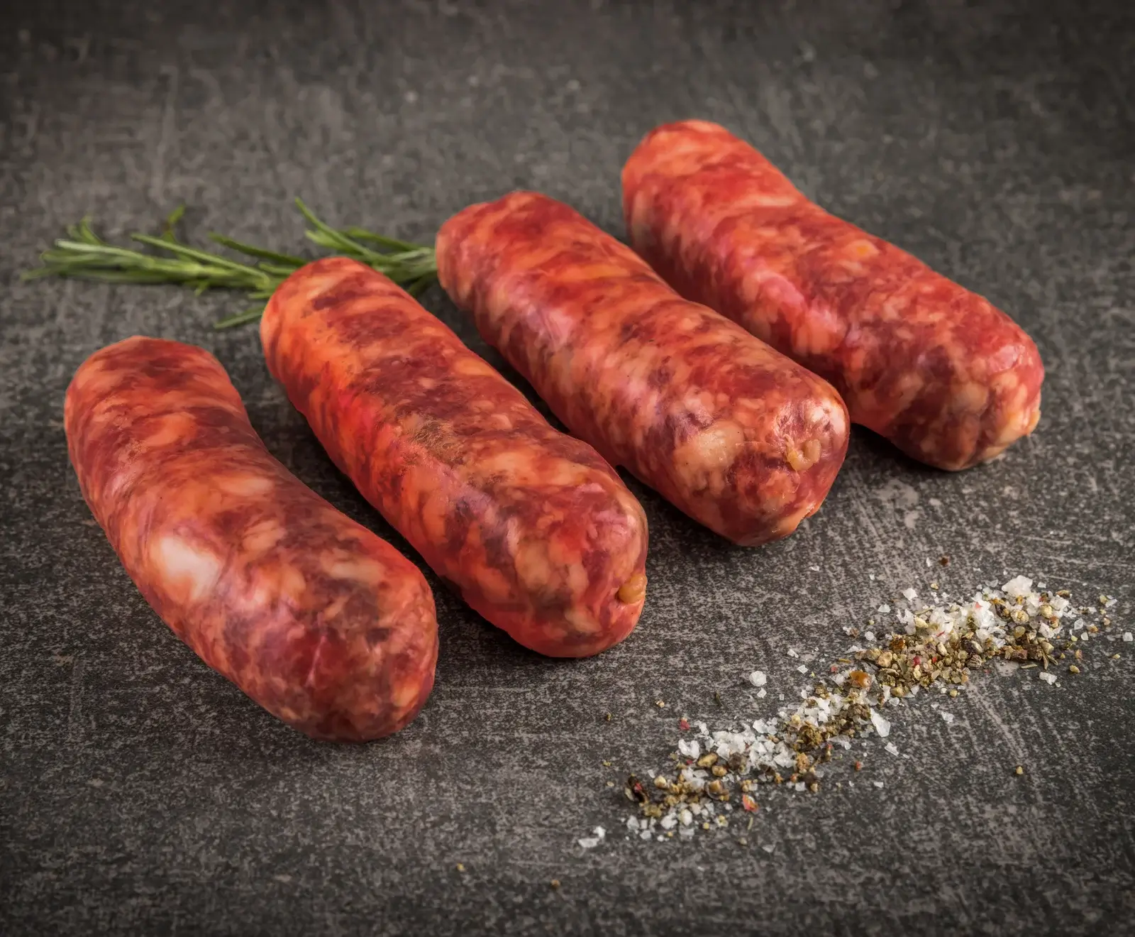 Vier rohe, rötliche Würste liegen auf einer dunklen Steinplatte. Im Hintergrund ist ein Rosmarinzweig, im Vordergrund grobes Salz und Pfeffer verstreut.