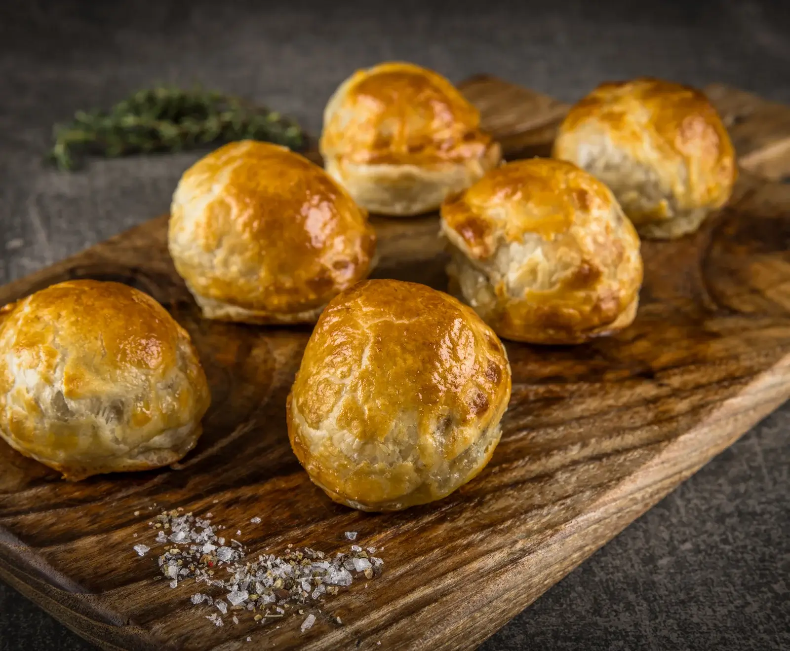 Sechs goldbraune, runde Blätterteigpasteten auf einem rustikalen Holzbrett, garniert mit grobem Salz, Pfeffer und einem Zweig Thymian im Hintergrund.