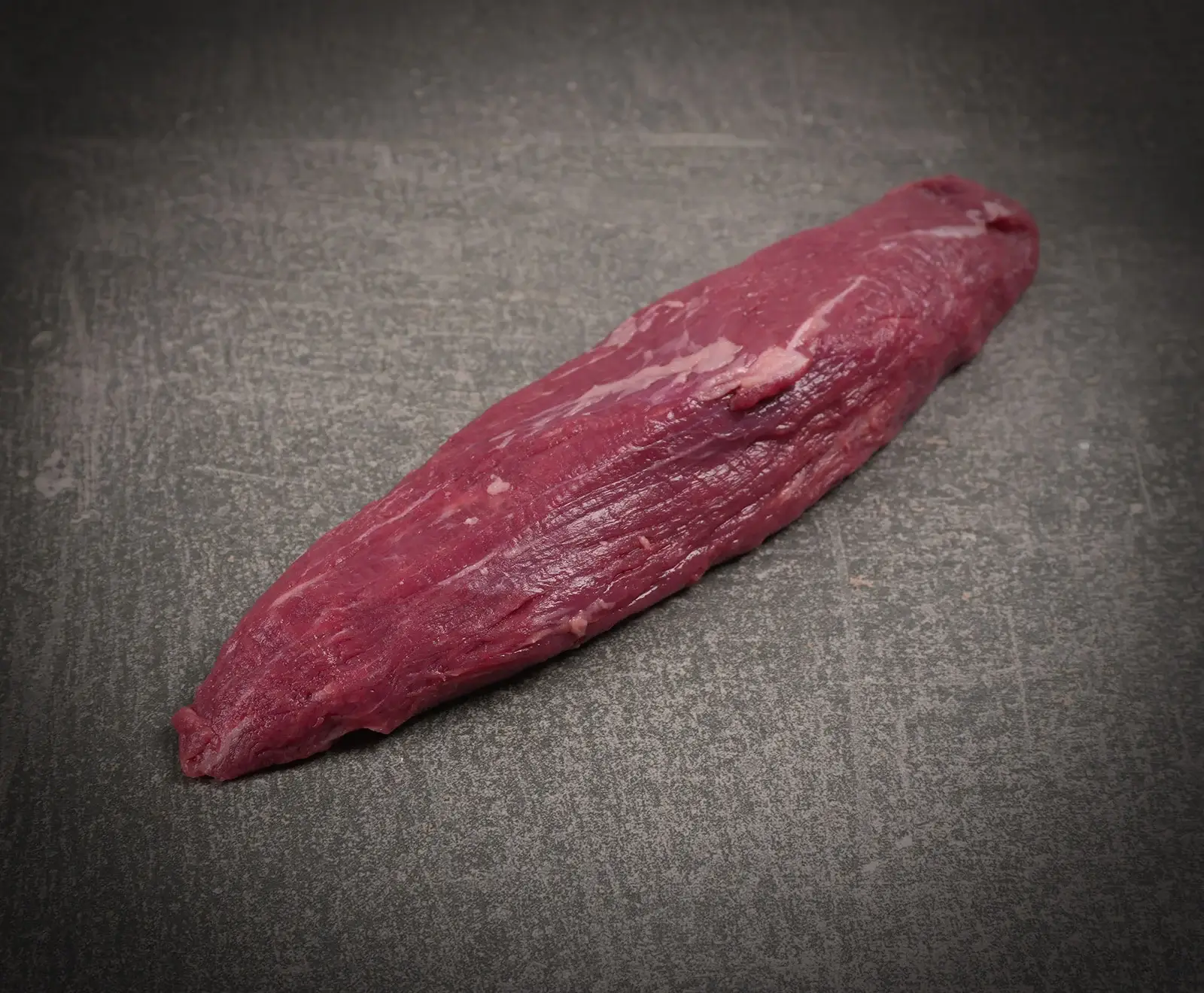 Ein längliches, rohes Stück dunkelrotes Fleisch, vermutlich ein Rinderfilet oder eine Lende, liegt diagonal auf einer dunklen, strukturierten Oberfläche. Es zeigt leichte Marmorierung.