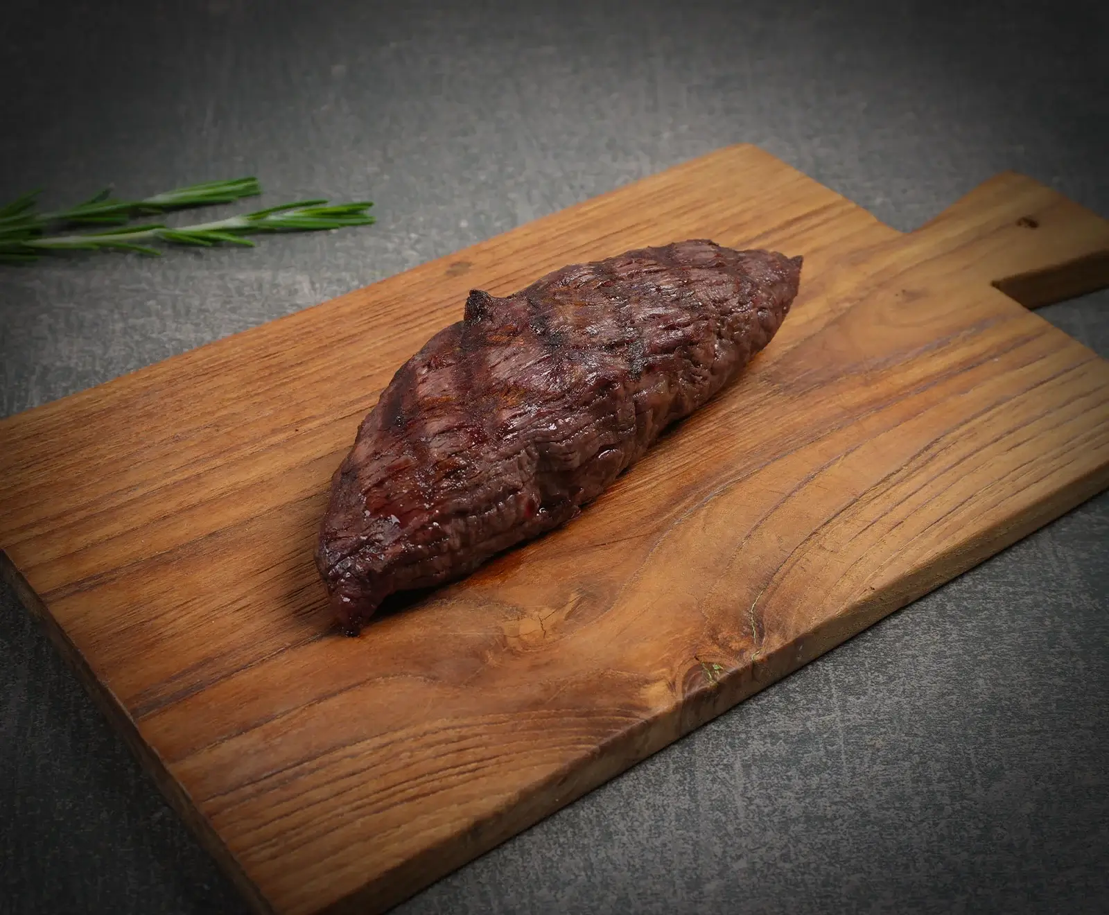 Ein gegrilltes Stück Fleisch, wahrscheinlich ein Steak, liegt auf einem rustikalen Holzschneidebrett. Im Hintergrund sind zwei Rosmarinzweige auf einer dunklen Oberfläche zu sehen.