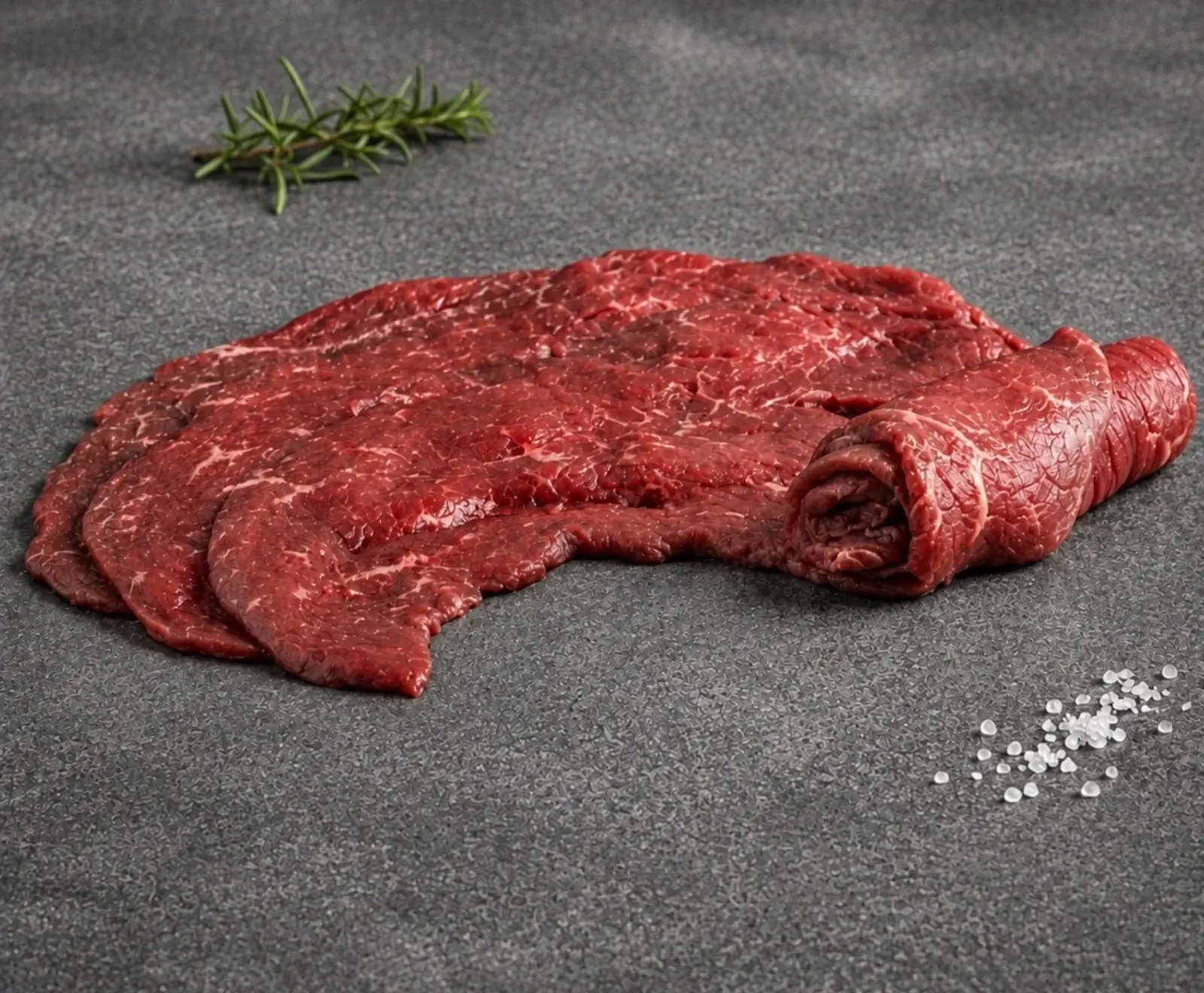 Rohes, dünn geschnittenes Rindfleisch, teilweise gerollt, auf dunklem Untergrund mit einem Rosmarinzweig und Meersalzflocken.
