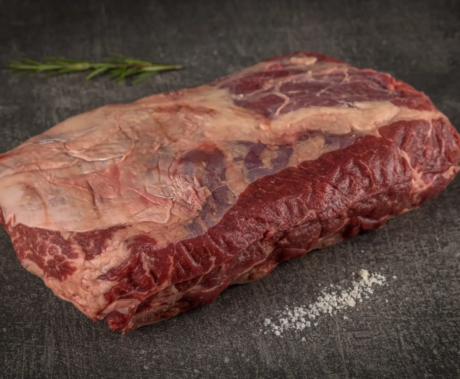 Ein großes, rohes Stück Rindfleisch mit Fettauflage und Marmorierung liegt auf einem dunklen Untergrund. Im Hintergrund ist ein Rosmarinzweig, im Vordergrund grobes Salz zu sehen.