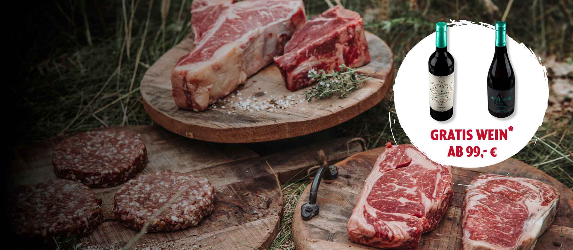 Verschiedene rohe Fleischstücke (Steaks, Burger) auf rustikalen Holzbrettern in natürlicher Umgebung. Eine Werbeaktion bietet zwei Flaschen Gratis Wein ab einem Einkaufswert von 99,- €.