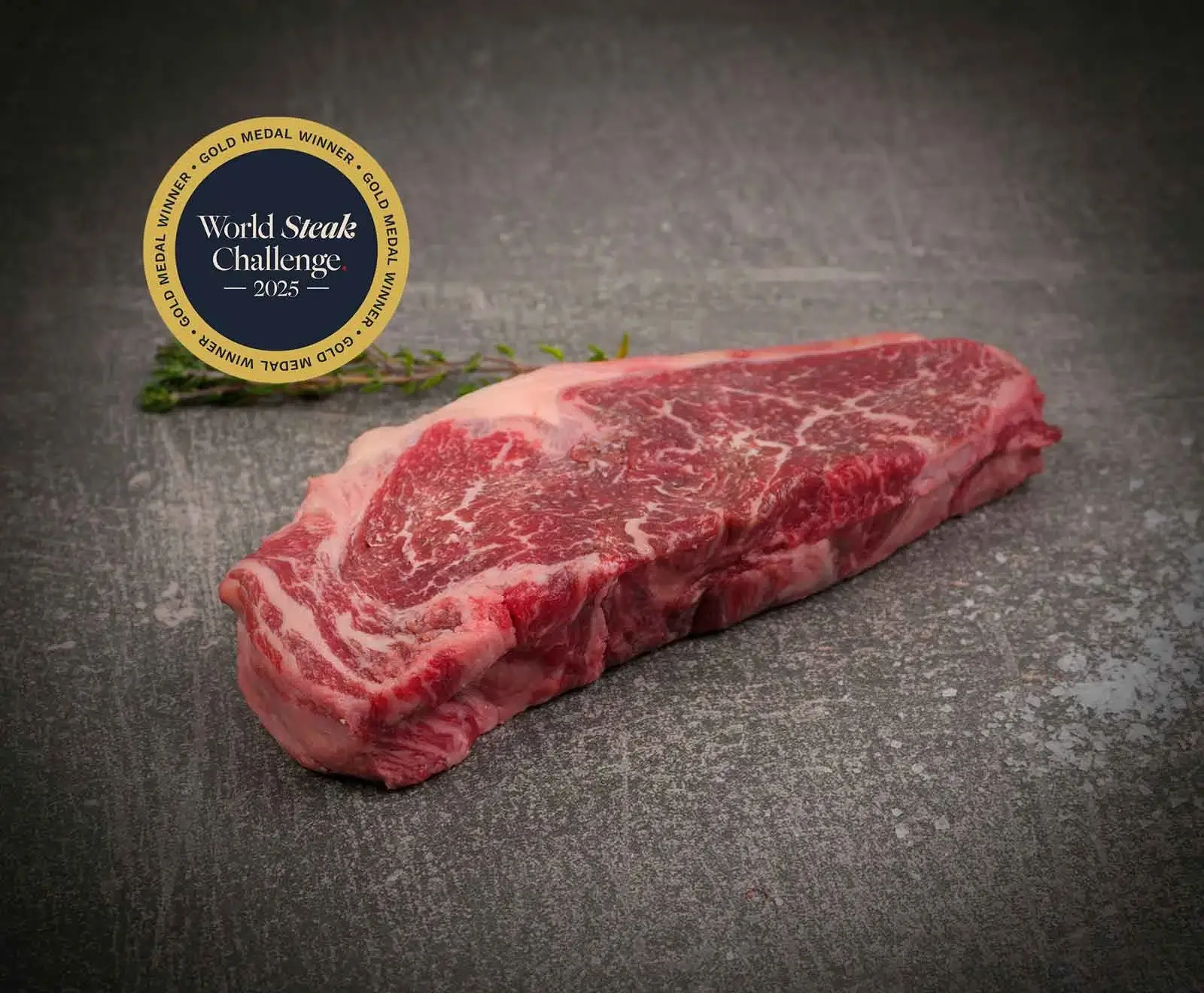 Ein rohes, gut marmoriertes Steak auf dunklem, strukturiertem Untergrund, mit einem Thymianzweig im Hintergrund. Oben links ein goldener Aufkleber: "World Steak Challenge 2023 Gold Medal Winner".
