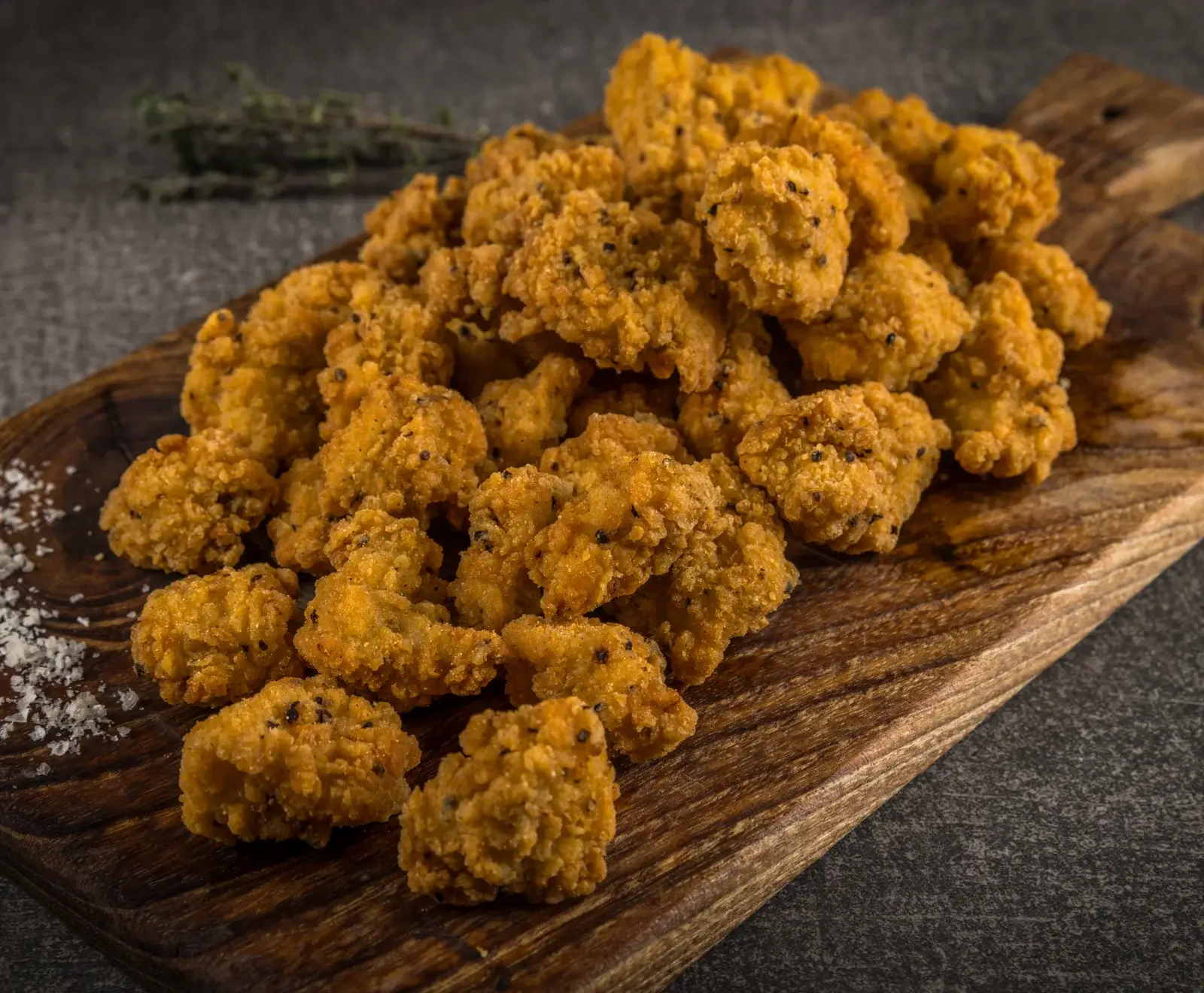 Ein Haufen goldbrauner, knuspriger Hähnchen-Popcorn auf einem rustikalen Holzbrett, mit grobem Salz und einem Kräuterzweig im unscharfen Hintergrund.