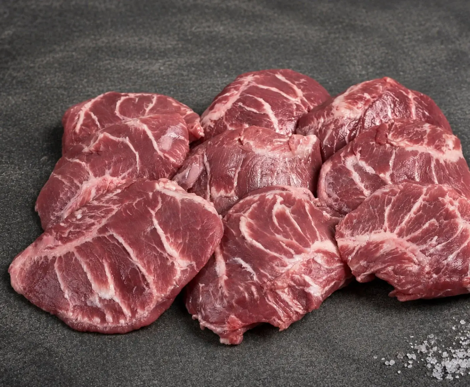 Mehrere rohe, rot-rosa Schweinenackensteaks mit deutlicher Marmorierung liegen auf einer dunklen, strukturierten Oberfläche. Rechts unten ist eine Prise grobes Salz zu sehen.
