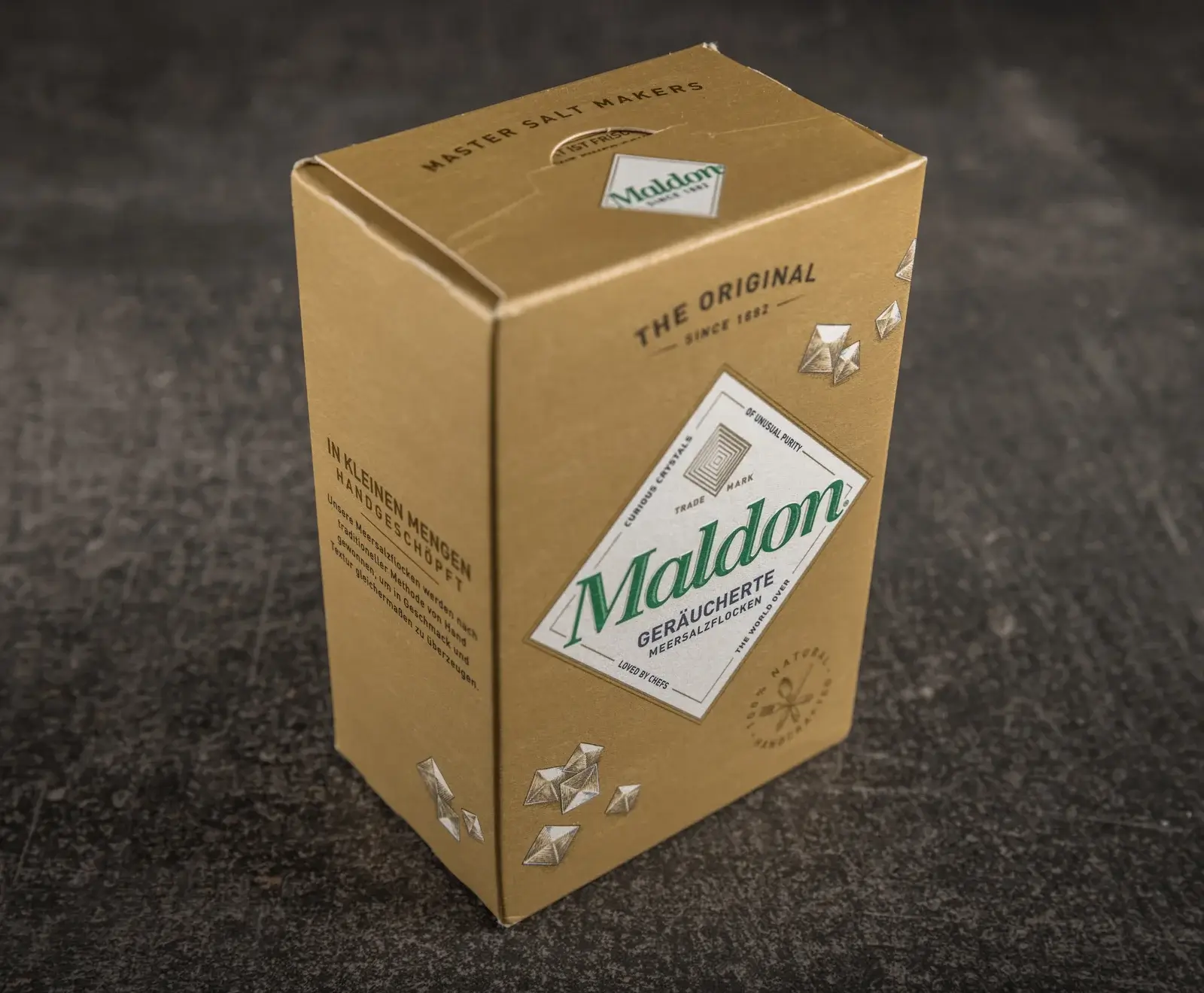 Ein brauner Karton Maldon Geräucherte Meersalzflocken steht schräg auf einem dunklen Untergrund. Die Vorderseite zeigt das grüne Maldon-Logo und Salz-Kristall-Illustrationen.