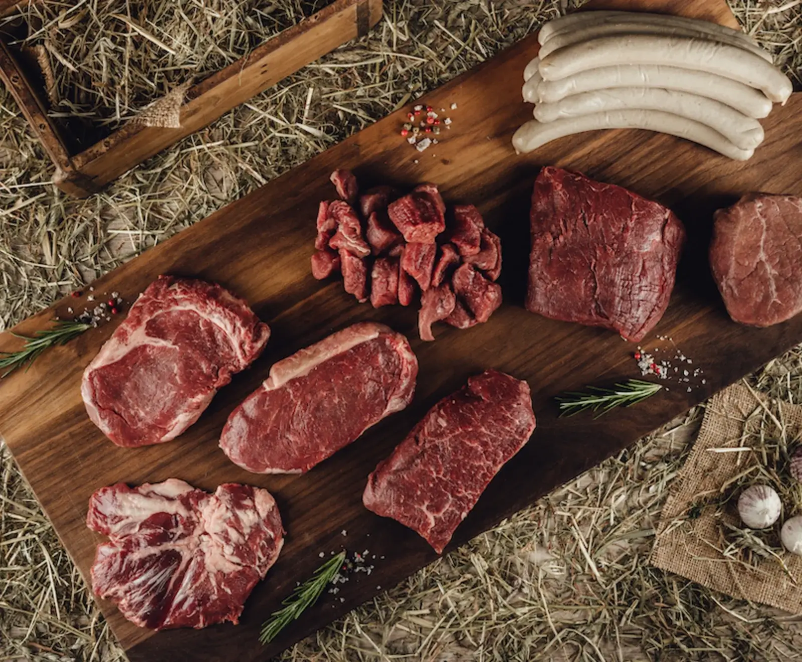 Verschiedene rohe Fleischstücke (Steaks, Würfel, Braten) und weiße Würste auf einem Holzbrett, umgeben von Heu. Garniert mit Rosmarin, Salz und Pfeffer.