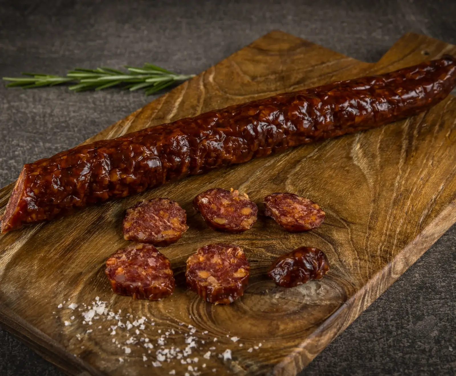Eine glänzende, dunkelrote Wurst liegt diagonal auf einem Holzschneidebrett. Daneben sind mehrere dicke Scheiben der Wurst, grobes Salz und ein Rosmarinzweig auf dunklem Hintergrund zu sehen.
