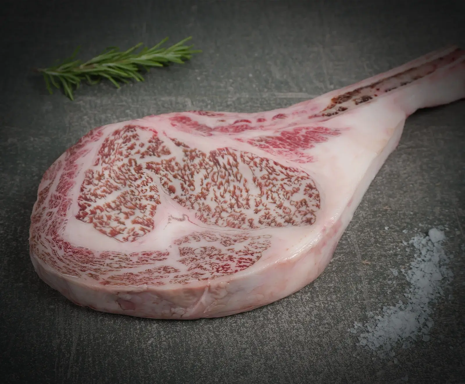 Ein rohes, stark marmoriertes Tomahawk-Steak liegt auf einer dunklen Oberfläche. Ein Rosmarinzweig und grobes Salz sind daneben platziert. Das Fleisch zeigt eine intensive Fettmarmorierung.