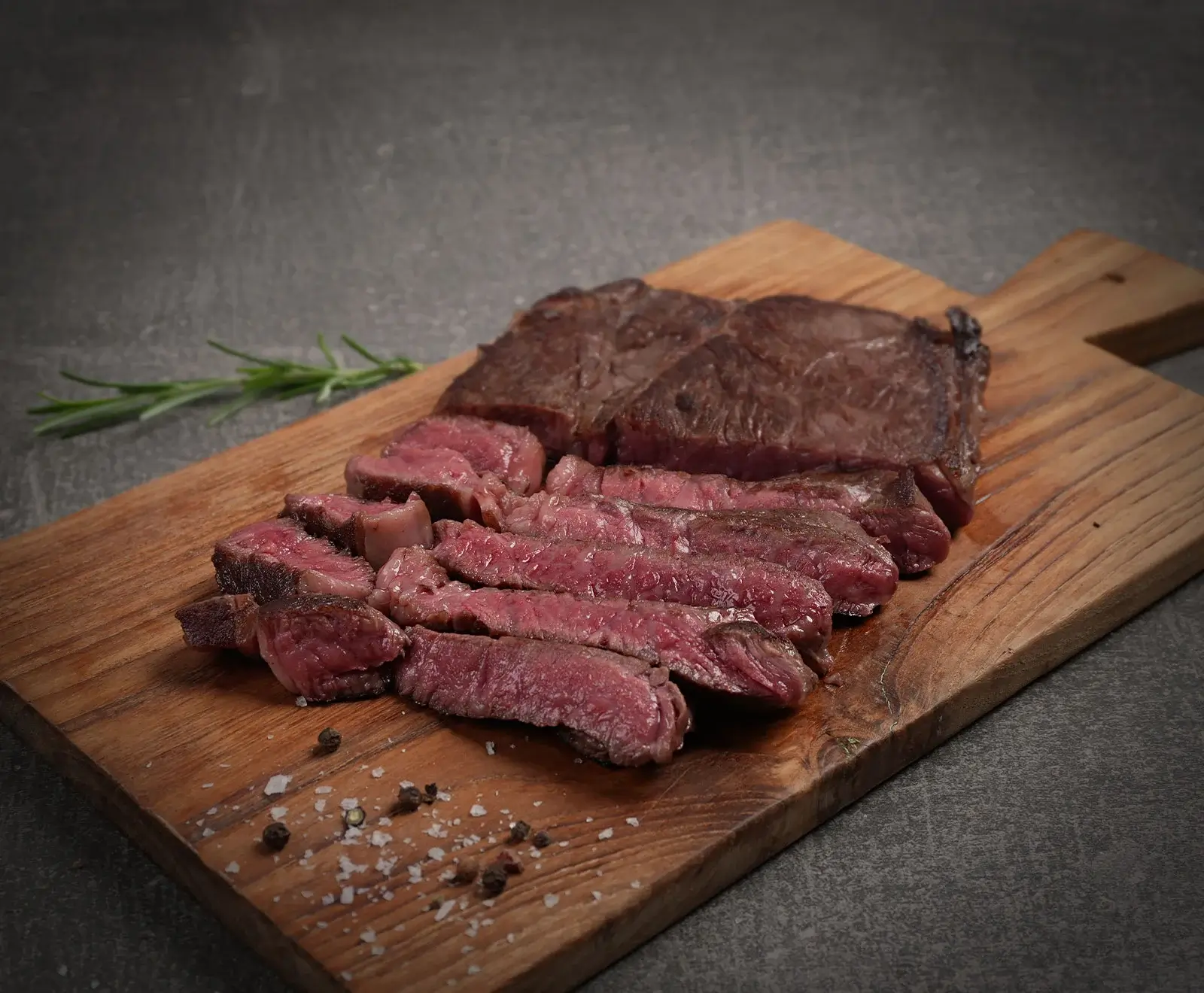 Ein aufgeschnittenes, medium-rare gegartes Steak auf einem Holzschneidebrett. Daneben liegen grobes Salz, schwarze Pfefferkörner und ein Rosmarinzweig auf dunklem Untergrund.