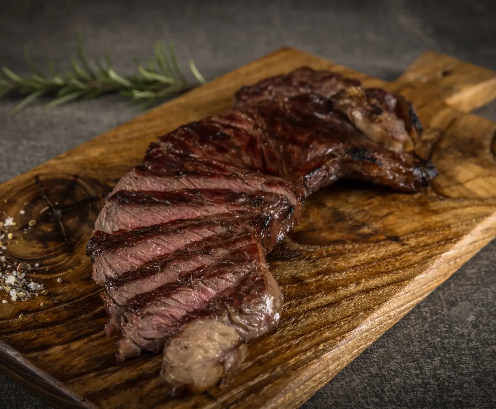 Ein saftiges, gegrilltes Rindersteak, teilweise in Scheiben geschnitten, liegt auf einem Holzschneidebrett. Daneben sind Rosmarin und grobes Salz mit Pfeffer zu sehen.