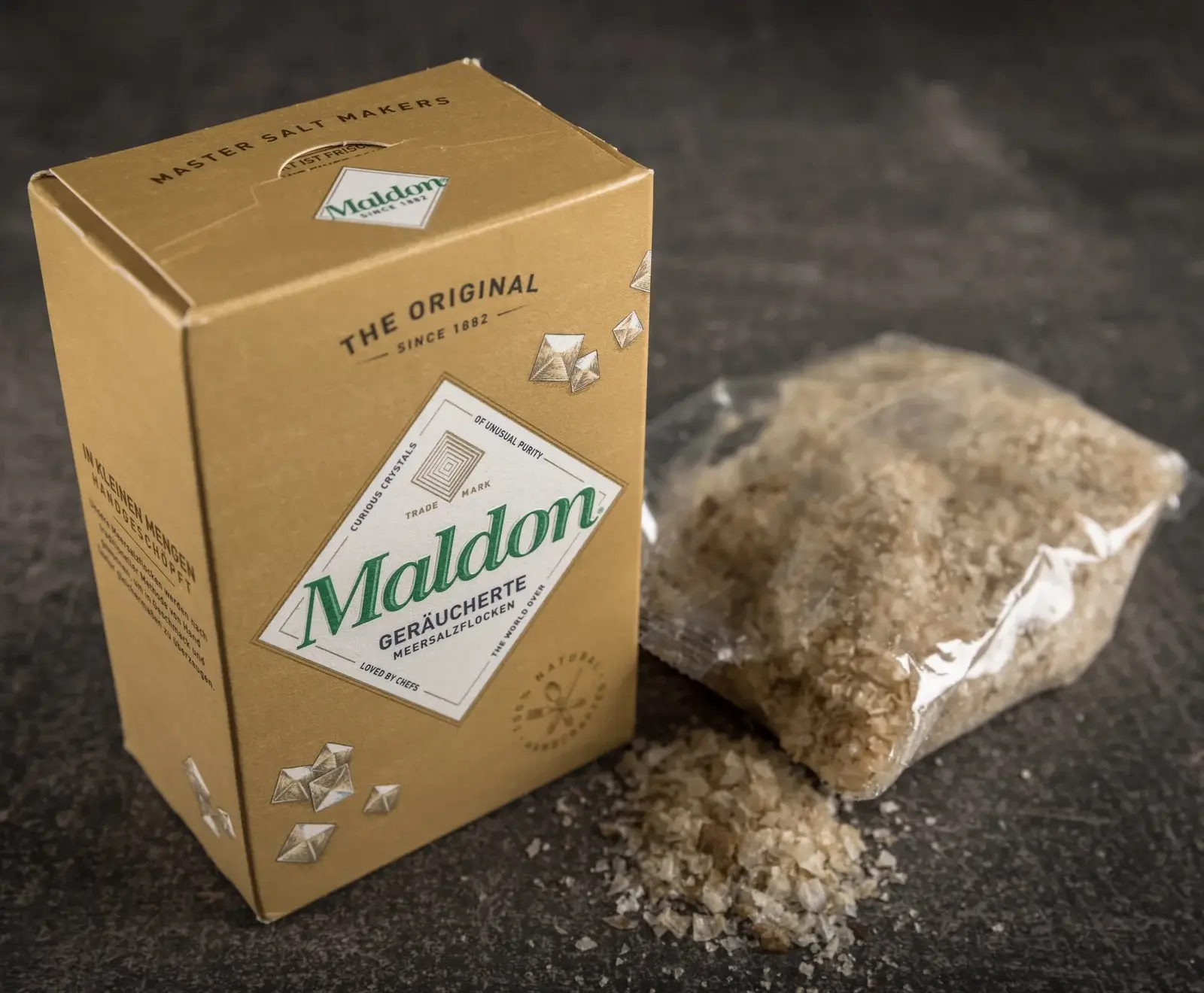 Eine goldbraune Maldon-Schachtel mit geräucherten Meersalzflocken steht neben einem offenen Plastikbeutel mit Salz, das teilweise auf einer dunklen Oberfläche verschüttet ist.