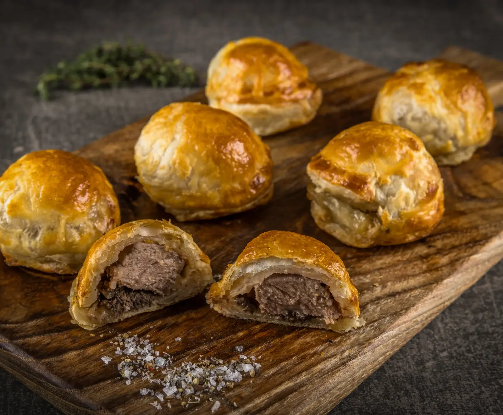 Sieben goldbraune Blätterteigtaschen mit Fleischfüllung auf einem Holzbrett. Zwei sind halbiert und zeigen rosa Fleisch und eine dunklere Schicht, bestreut mit grobem Salz.