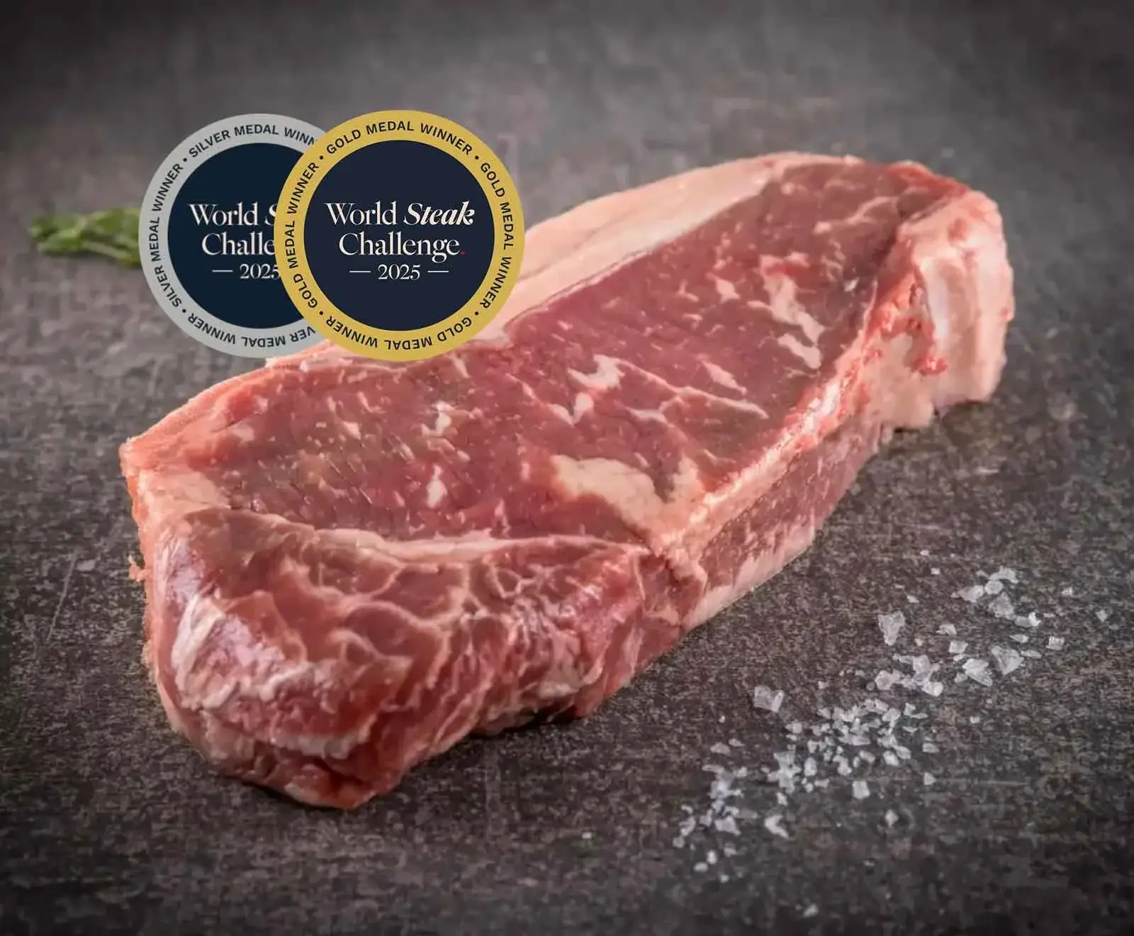 Rohes, marmoriertes Steak auf dunklem Untergrund mit grobem Salz und zwei Medaillen "World Steak Challenge 2025" (Gold und Silber).