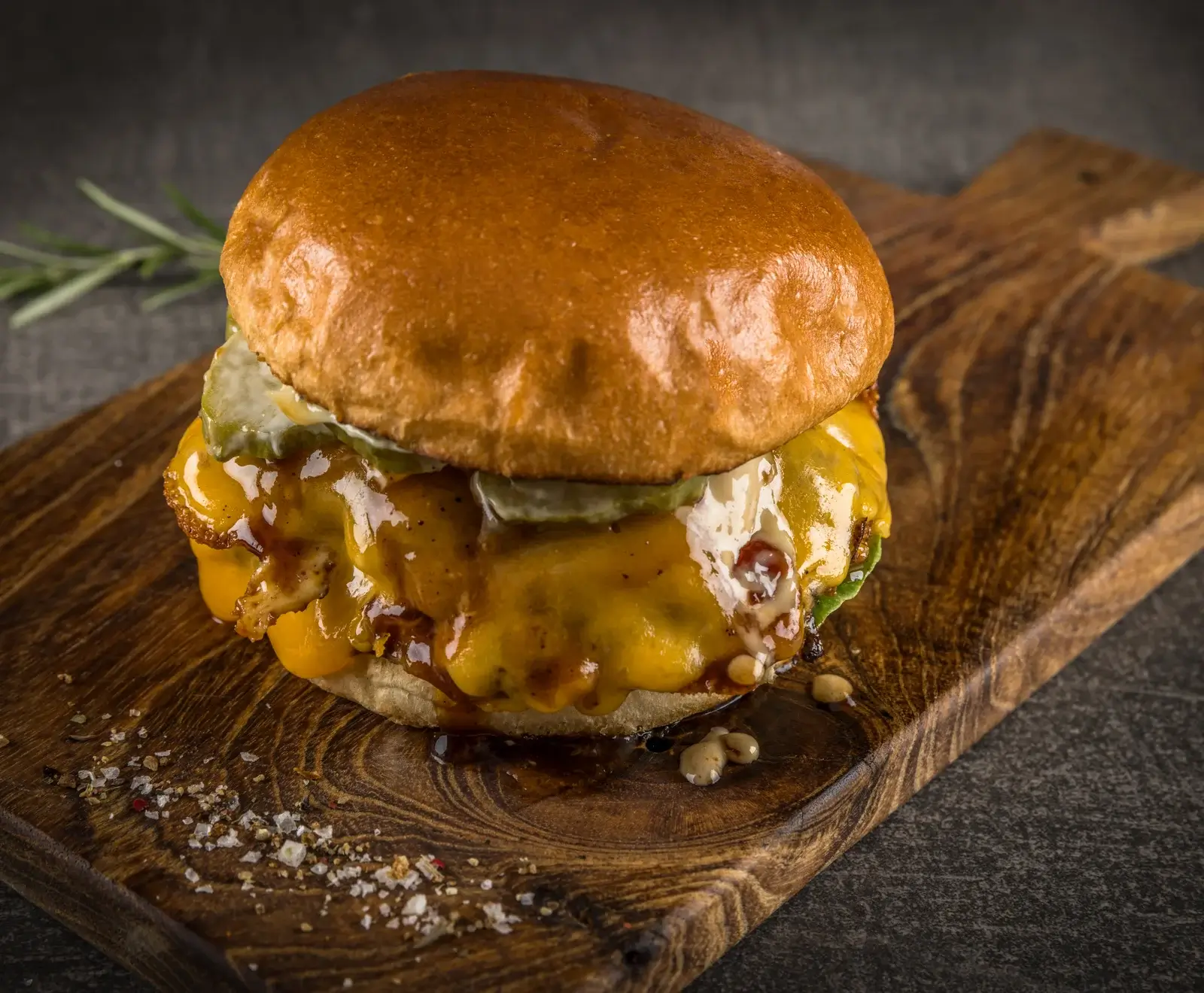 Nahaufnahme eines saftigen Burgers mit goldbraunem Brötchen, viel geschmolzenem Käse, Gurken und dunkler Soße, serviert auf einem rustikalen Holzbrett mit Salz und Pfeffer.