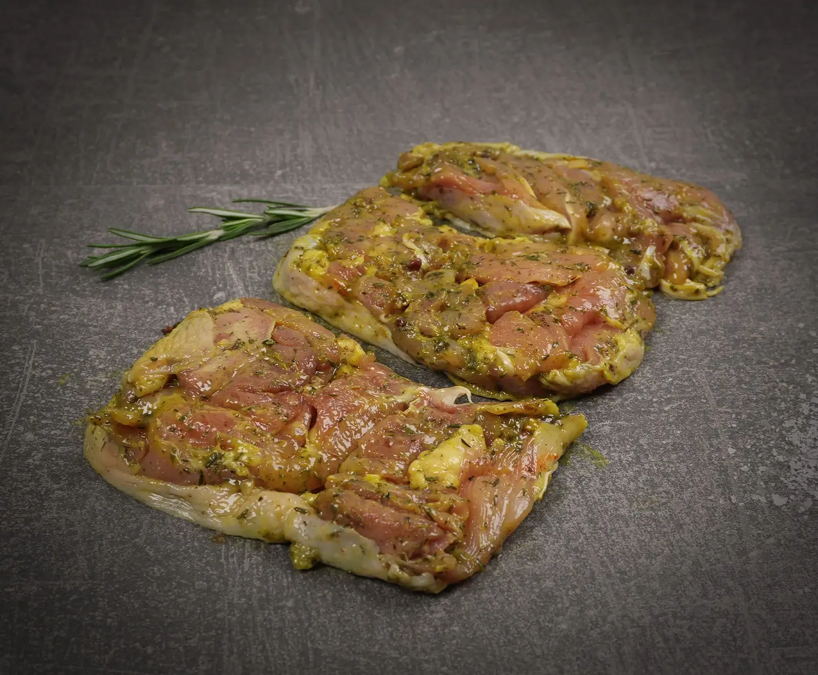 Drei Stücke rohes Hähnchenfleisch, mariniert in einer gelb-grünlichen Kräutermischung, liegen auf einem dunkelgrauen Untergrund. Daneben ist ein frischer Rosmarinzweig platziert.