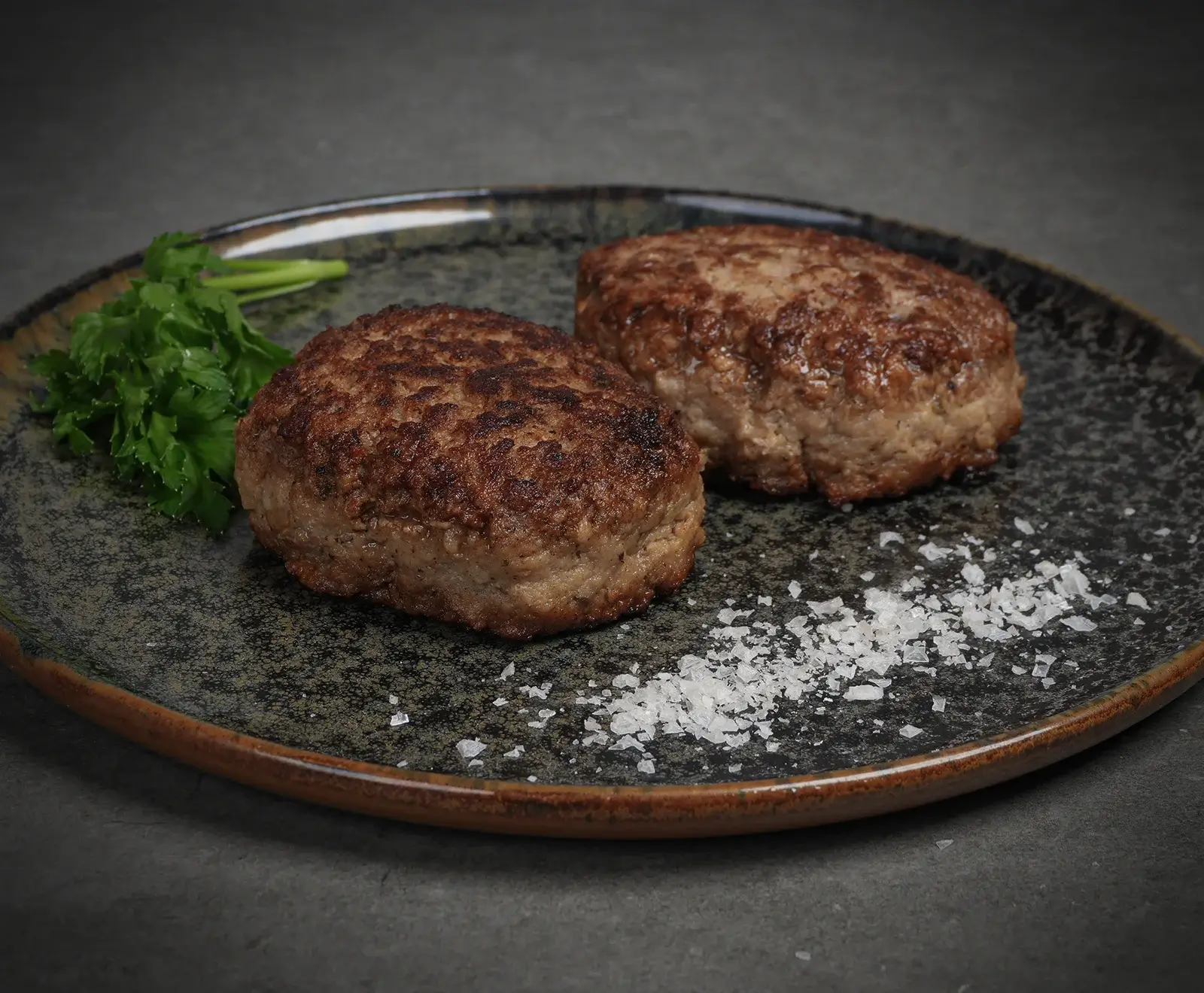 Zwei goldbraun gebratene Fleischpflanzerl auf einem dunklen, gesprenkelten Teller, garniert mit frischer Petersilie und groben Meersalzkristallen.
