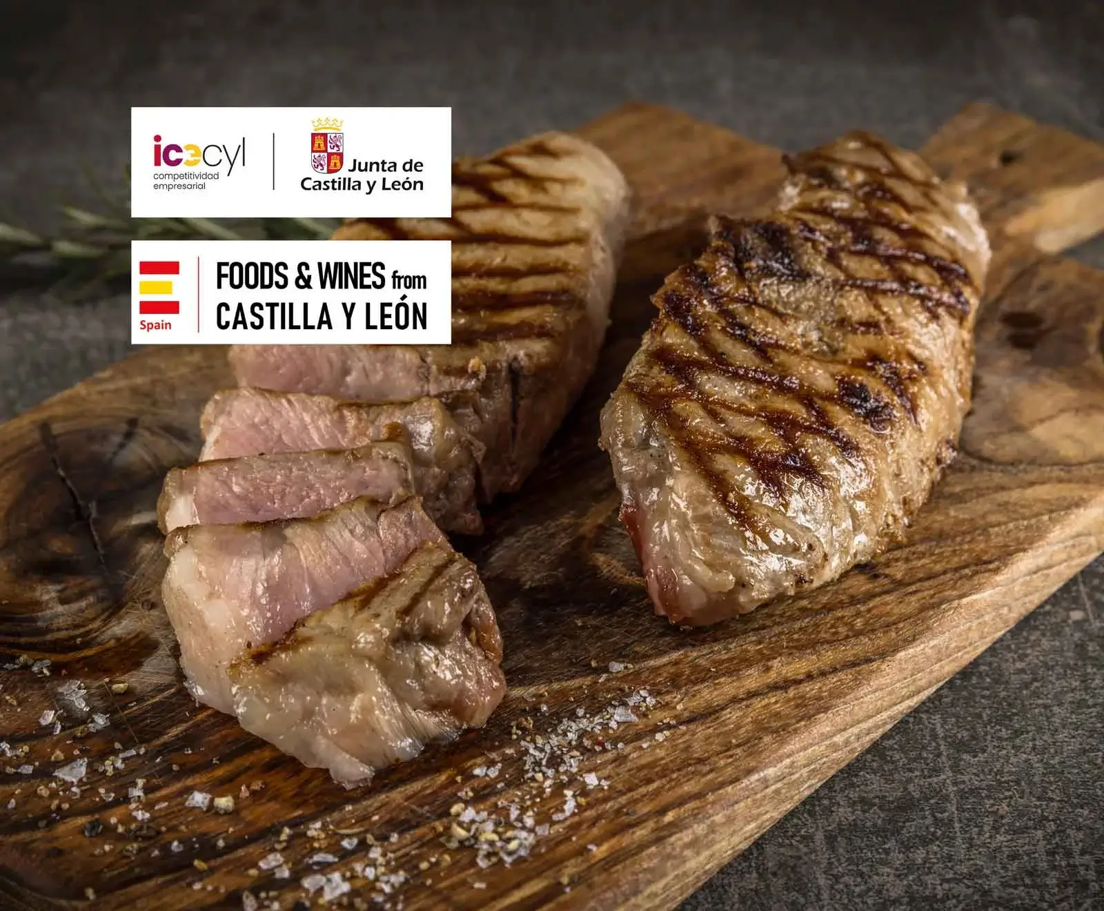 Zwei gegrillte Steaks auf einem Holzschneidebrett, eines geschnitten, mit grobem Salz und Pfeffer bestreut. Oben links Logos von "Junta de Castilla y León" und "FOODS & WINES from CASTILLA Y LEÓN Spain".