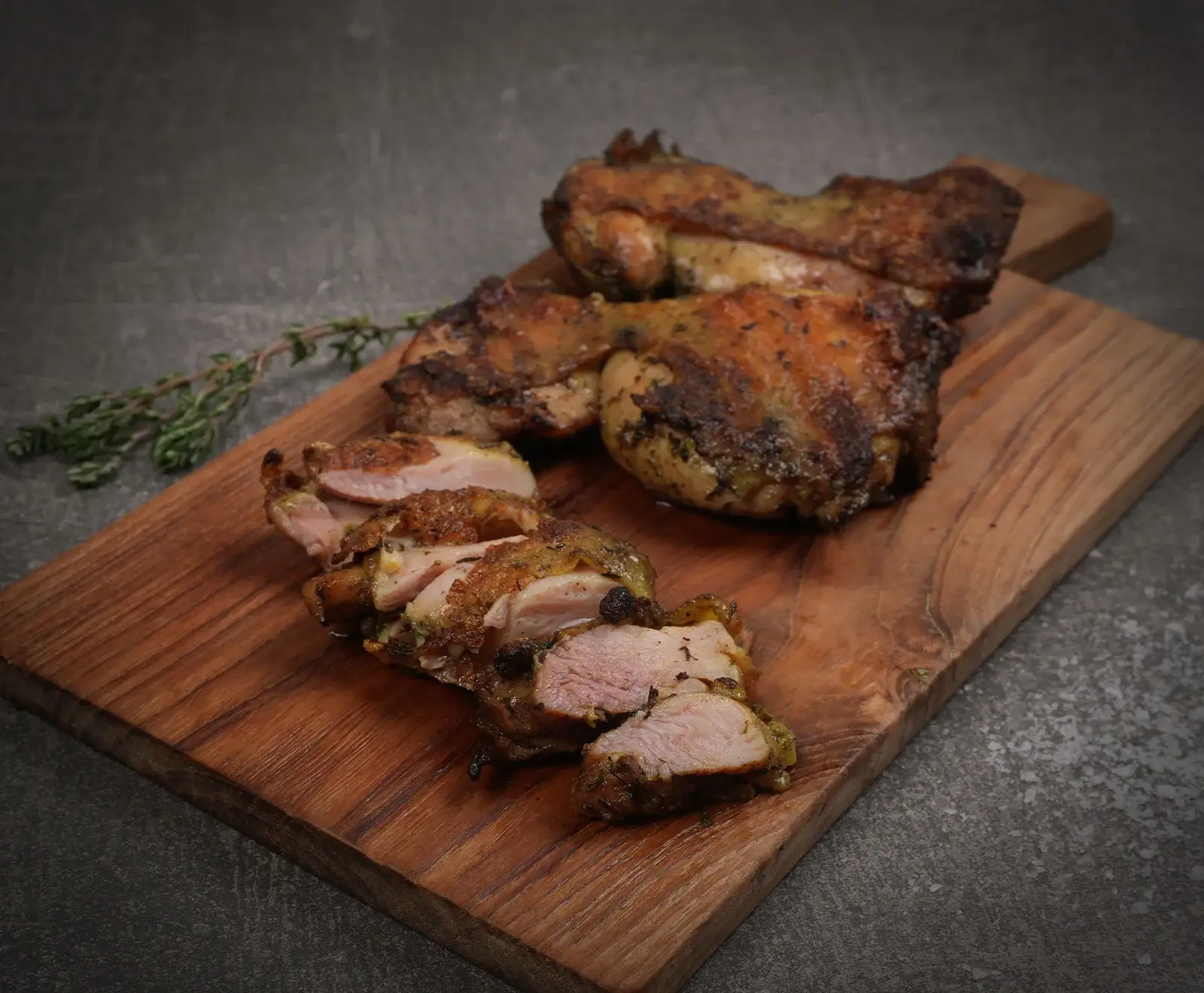 Drei knusprig gebratene Hähnchenteile, eines davon in Scheiben geschnitten, liegen auf einem rustikalen Holzschneidebrett. Ein Zweig Thymian ist ebenfalls zu sehen.