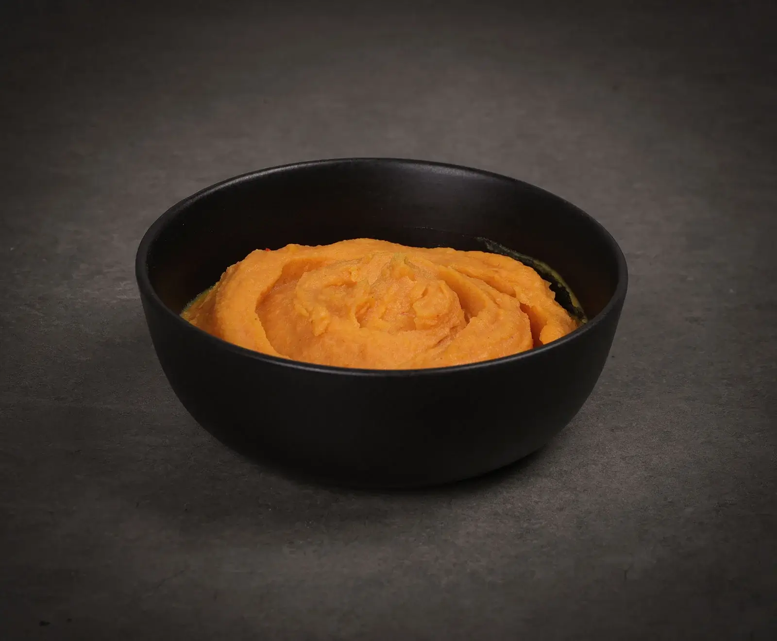 Nahaufnahme einer schwarzen Schale mit cremigem, leuchtend orangefarbenem Püree, das in der Mitte eine leichte Spirale bildet, auf einer dunklen Oberfläche.