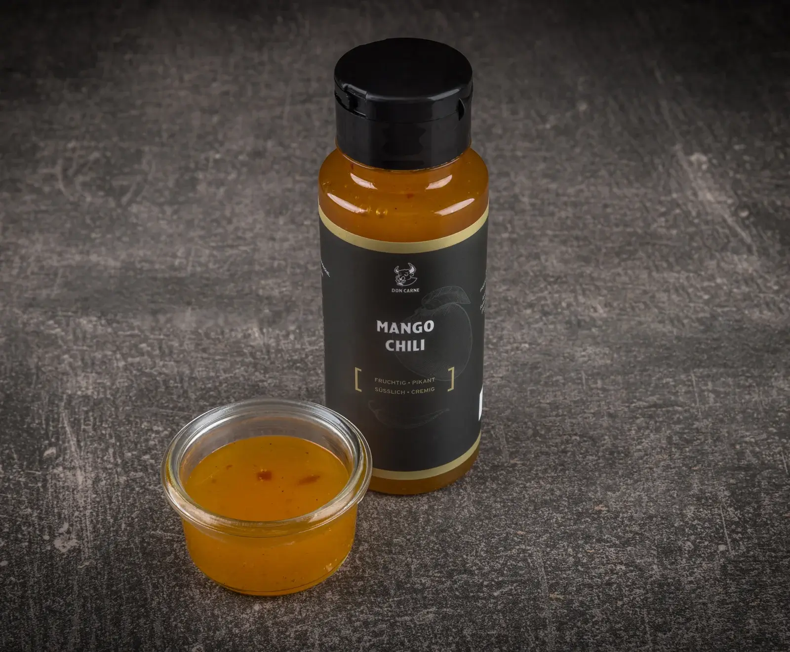 Eine schwarze Flasche Don Carne Mango Chili Sauce und eine kleine Glasschale mit der orangefarbenen Sauce stehen auf dunklem, strukturiertem Untergrund. Die Flasche ist mit "Fruchtig • Pikant Süsslich • Cremig" beschriftet.