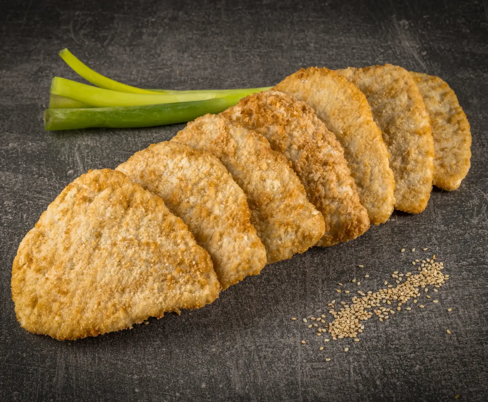 Sieben panierte, dreieckige Lebensmittelstücke liegen diagonal auf einer dunklen Oberfläche. Oben links sind zwei Lauchzwiebeln, unten rechts eine kleine Menge Sesamsamen.