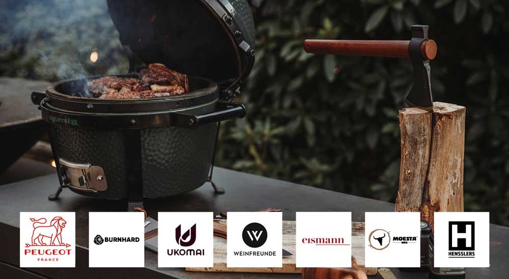 Ein Big Green Egg Kamado-Grill mit rauchendem Fleisch, daneben eine Axt in einem Holzscheit. Darunter Logos von Peugeot, Burnhard, Ukomai, Weinfreunde, Eismann, Moesta BBQ und Hensslers.