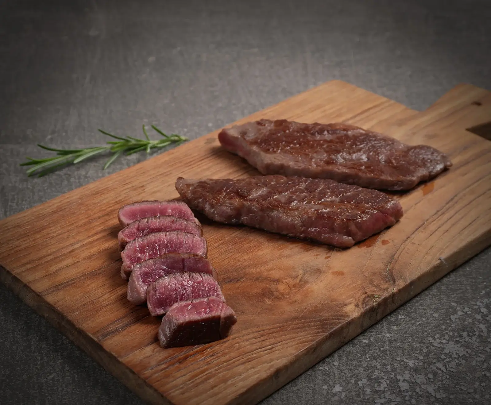 Zwei gebratene Steaks und ein in Scheiben geschnittenes Steak, medium-rare, auf einem Holzschneidebrett mit einem Rosmarinzweig auf dunklem Hintergrund.