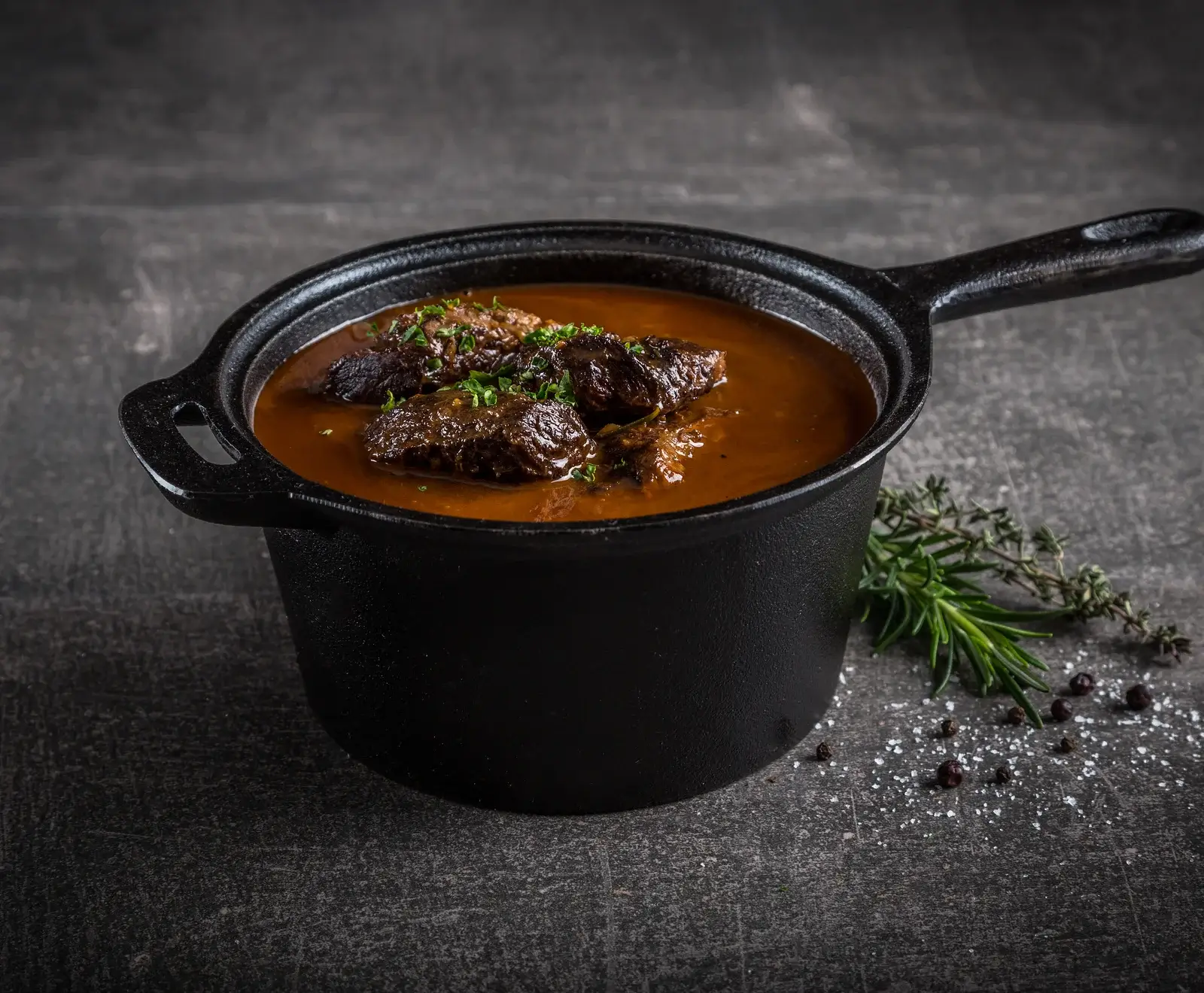 Ein schwarzer Gusseisentopf mit einem deftigen Schmorgericht, bestehend aus dunklem Fleisch in brauner Soße, garniert mit frischen Kräutern. Daneben liegen Rosmarin, Thymian, Pfeffer und Salz auf dunklem Untergrund.