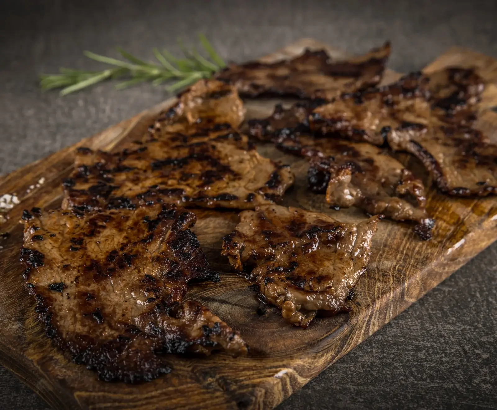 Dünn geschnittenes, gegrilltes Fleisch mit deutlichen Grillstreifen und glänzender Oberfläche, appetitlich auf einem rustikalen Holzbrett angerichtet. Ein Rosmarinzweig liegt im Hintergrund.
