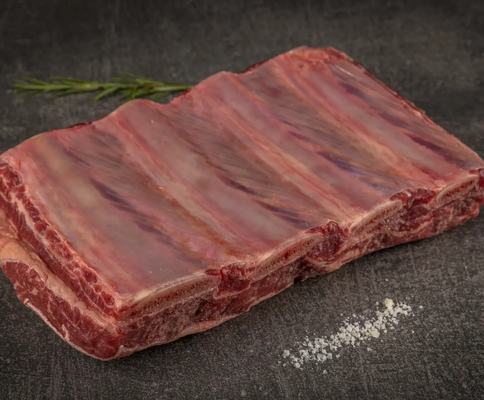Ein großes Stück rohe Rinderquerrippe mit sichtbaren Knochen und Fleisch, auf einer dunklen Oberfläche liegend. Im Hintergrund ein Rosmarinzweig, im Vordergrund grobes Salz.