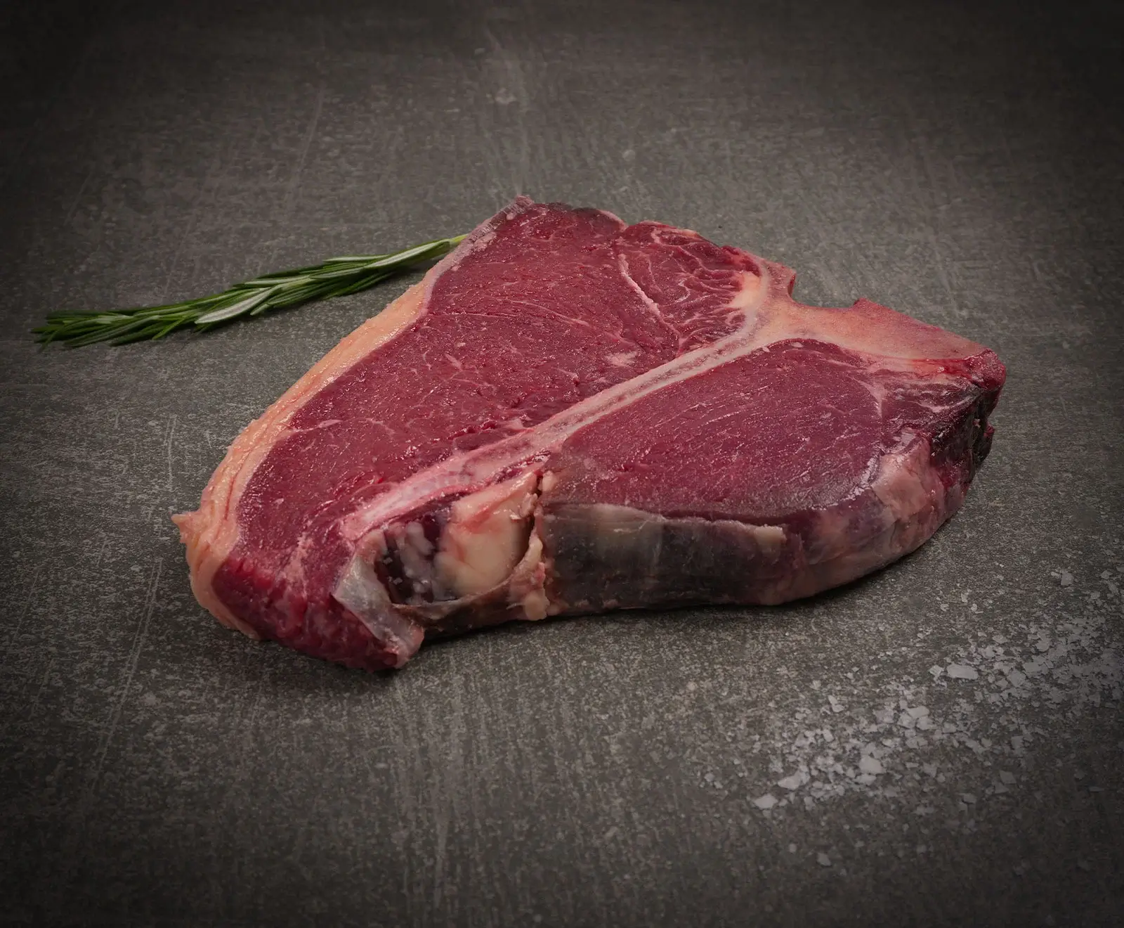 Ein rohes T-Bone Steak liegt auf einer dunklen Steinoberfläche. Links daneben befindet sich ein Rosmarinzweig, und im Vordergrund sind grobe Salzkristalle verstreut. Das Steak zeigt eine deutliche Marmorierung.