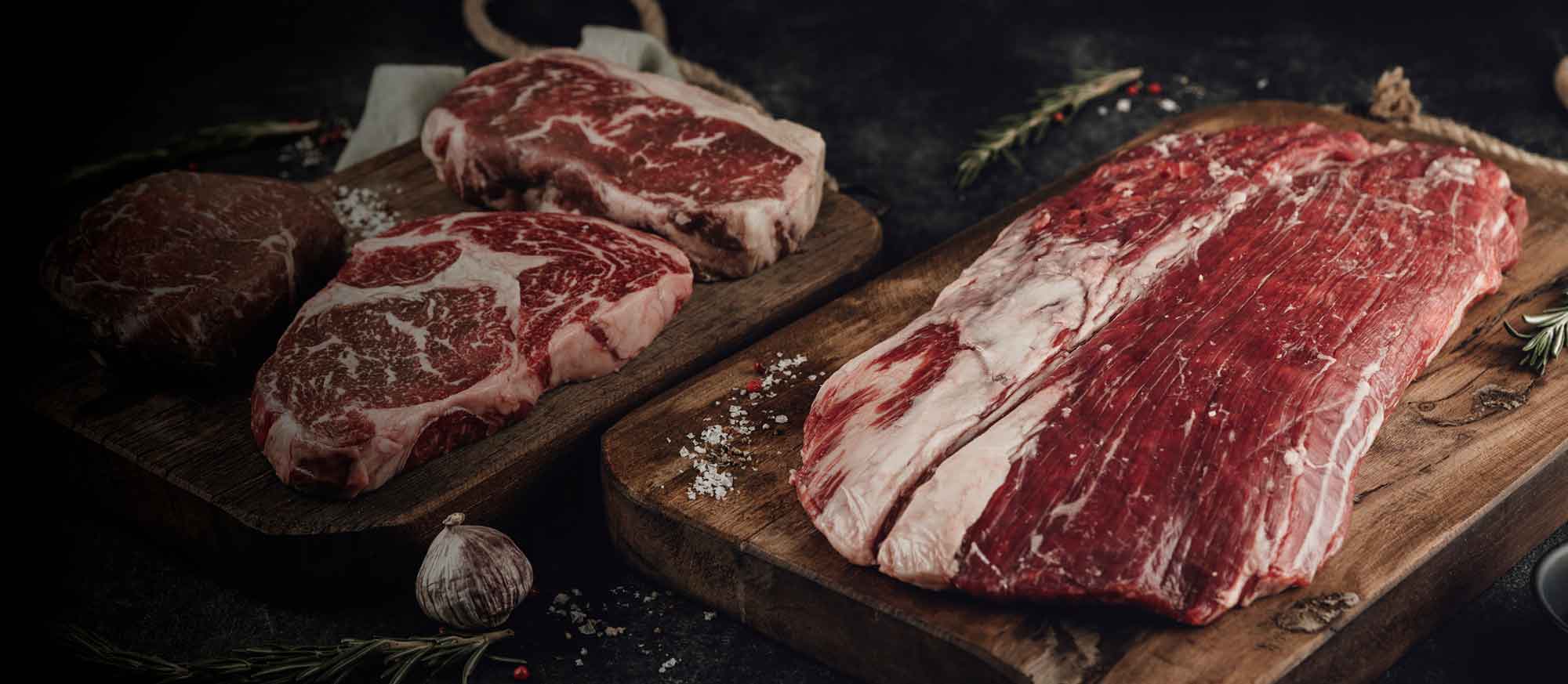 Drei rohe Rindfleischstücke auf zwei rustikalen Holzschneidebrettern. Links zwei marmorierte Steaks, rechts ein großes, flaches Stück Fleisch. Grobes Salz, Rosmarin und Knoblauch liegen daneben.