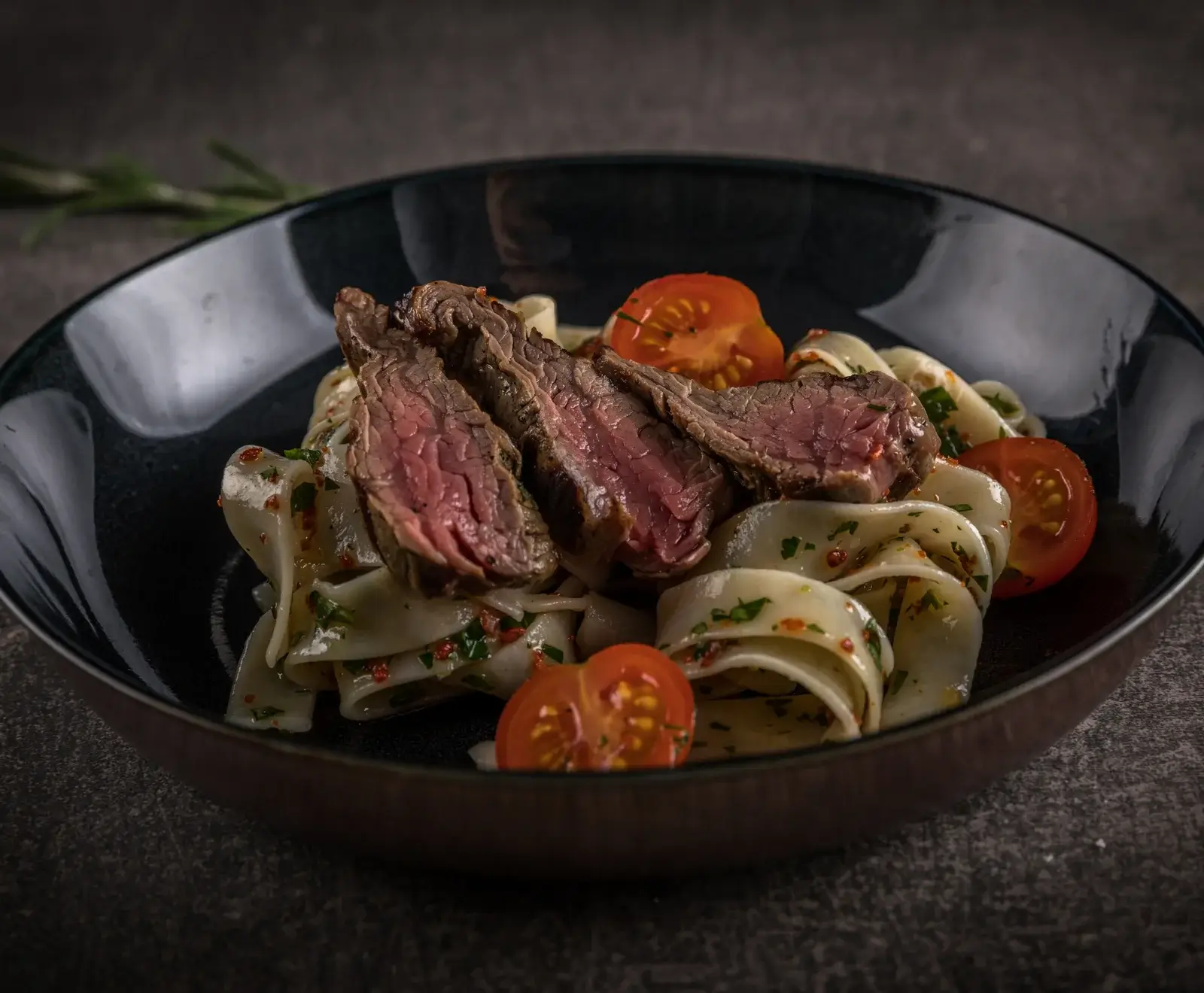Nahaufnahme eines dunklen Tellers mit drei Scheiben medium-rare Steak auf breiten Nudeln, garniert mit halbierten Kirschtomaten und Kräutern. Der Hintergrund ist dunkel und unscharf.