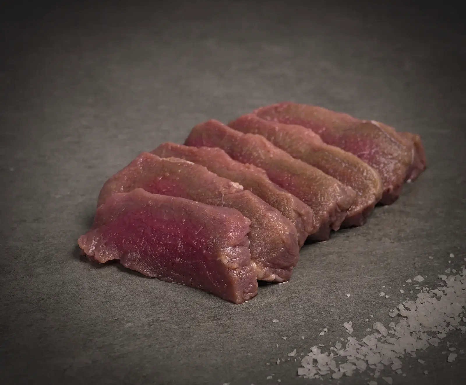Sieben Scheiben rohes, mageres Fleisch, möglicherweise Wild, liegen leicht überlappend auf einer dunklen Schieferplatte. Rechts unten sind grobe Meersalzflocken verstreut.