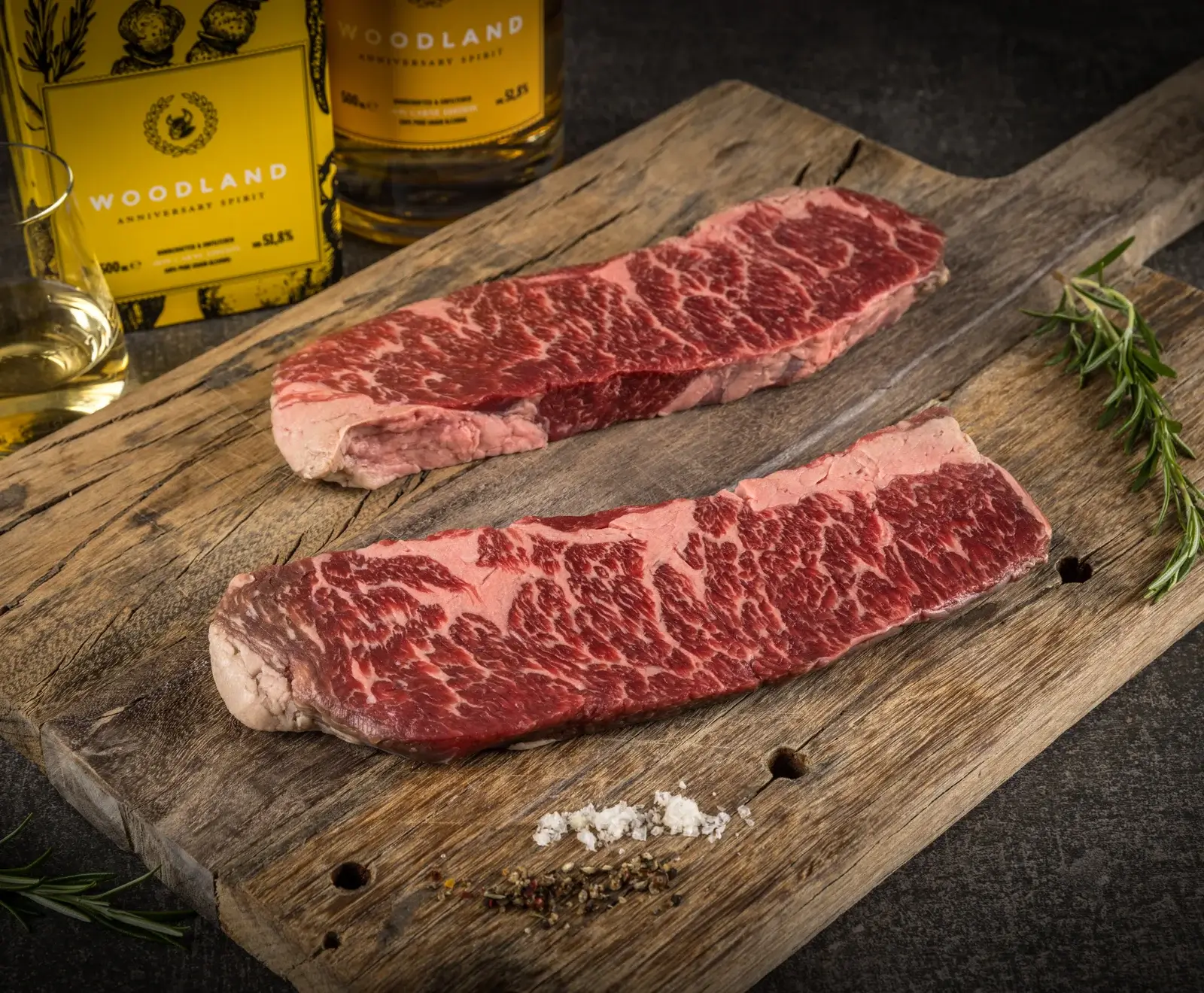 Zwei rohe, stark marmorierte Steaks liegen auf einem rustikalen Holzbrett, daneben grobes Salz, Pfeffer und Rosmarin. Im Hintergrund sind unscharf zwei Flaschen "WOODLAND ANNIVERSARY SPIRIT" und ein Glas mit goldener Flüssigkeit zu sehen.
