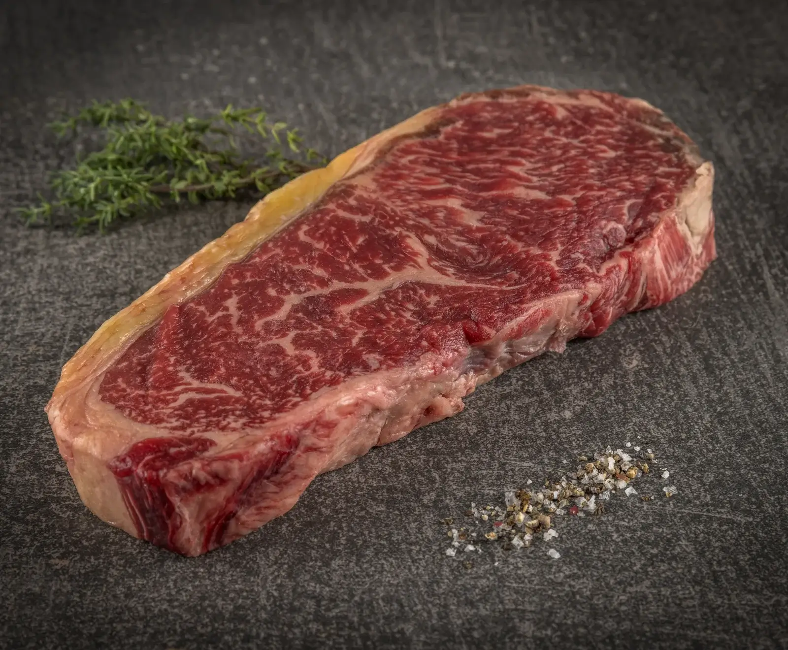 Ein rohes, stark marmoriertes Rindersteak mit Fettrand liegt auf einer dunklen Steinplatte. Daneben sind ein Zweig Thymian und eine Prise grobes Salz und Pfeffer zu sehen.