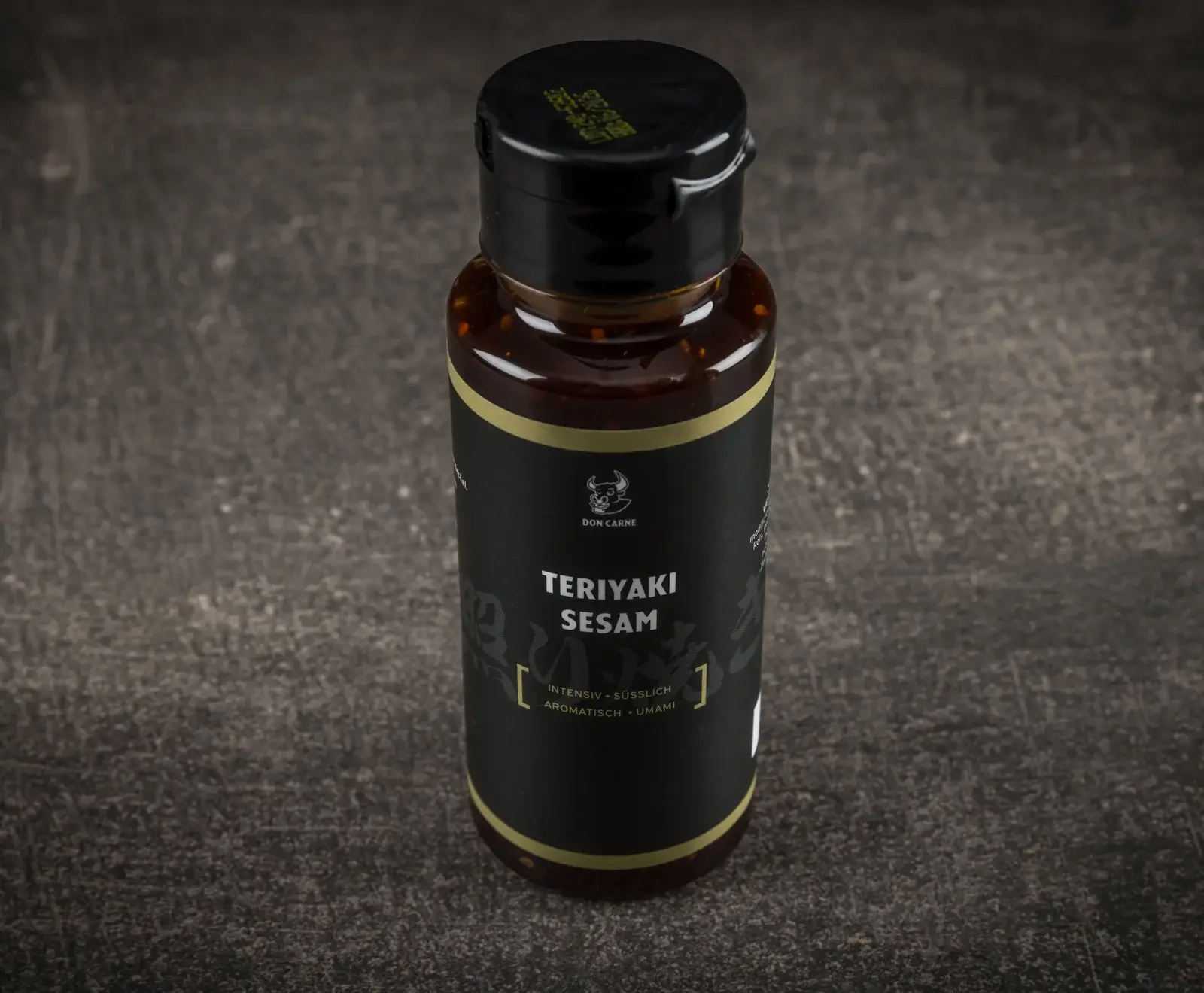 Eine Flasche Don Carne Teriyaki Sesam Sauce mit schwarzem Etikett, goldener Umrandung und schwarzem Klappdeckel, gefüllt mit dunkler Sauce, steht auf einem dunklen, strukturierten Untergrund.