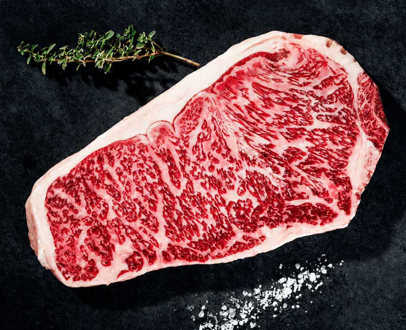 Ein rohes, stark marmoriertes Wagyu-Rindersteak liegt auf einer dunklen Oberfläche. Ein Zweig frischer Thymian ist oben links platziert, während grobes Meersalz unten rechts verstreut ist. Das Fleisch zeigt eine ausgeprägte, feine Fettzeichnung.