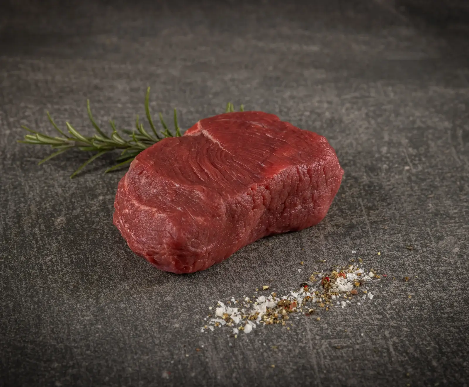Ein rohes, rotes Rinderfilet liegt auf einer dunklen Steinplatte, daneben ein Rosmarinzweig und eine Prise grobes Salz mit Pfeffer.
