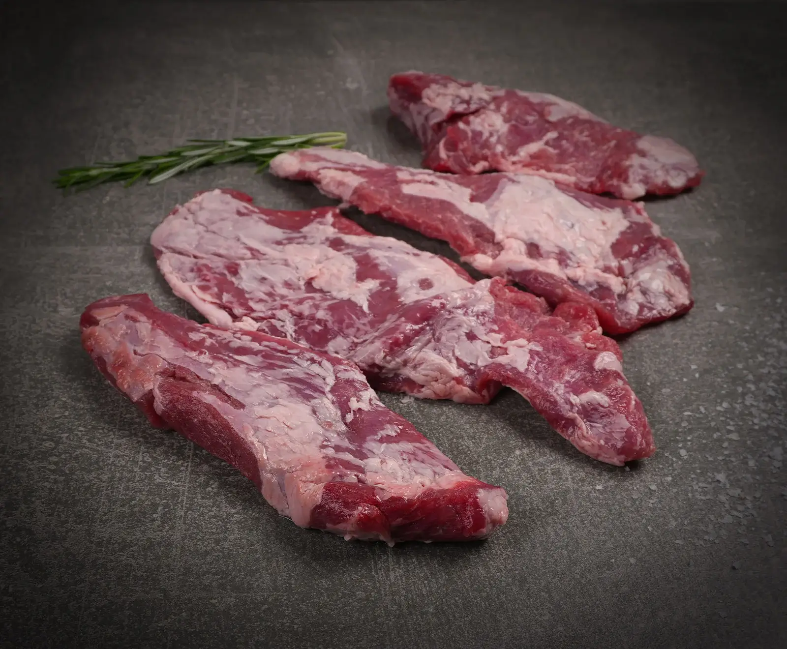 Vier Stücke rohes, mageres Fleisch mit etwas Fettauflage und Marmorierung liegen auf einer dunklen, strukturierten Oberfläche. Ein Rosmarinzweig ist im Hintergrund.